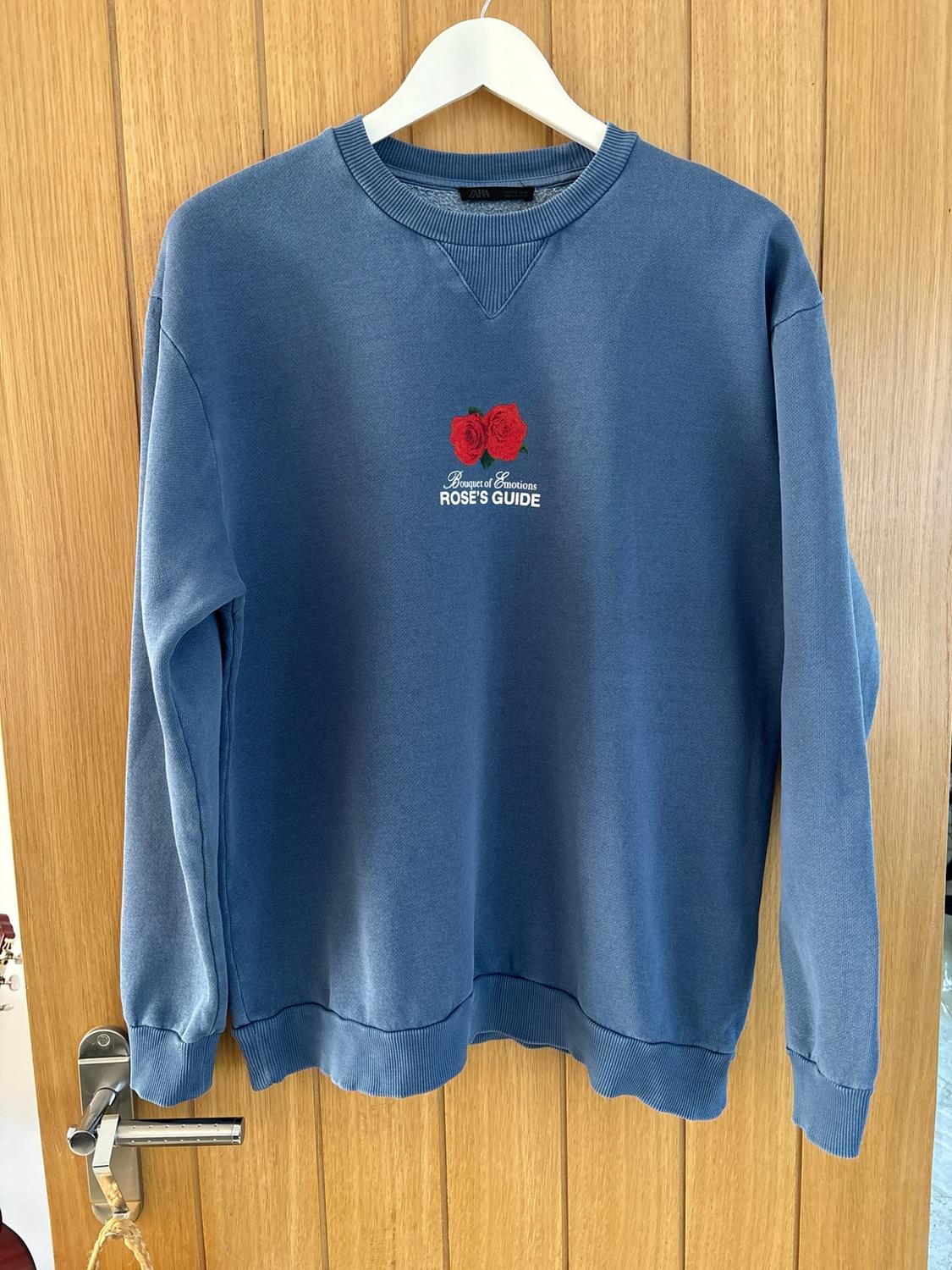 Blue sweatshirt hanging on a wooden door with two red roses and the text Bouquet of Emotions ROSE'S GUIDE in the centre.