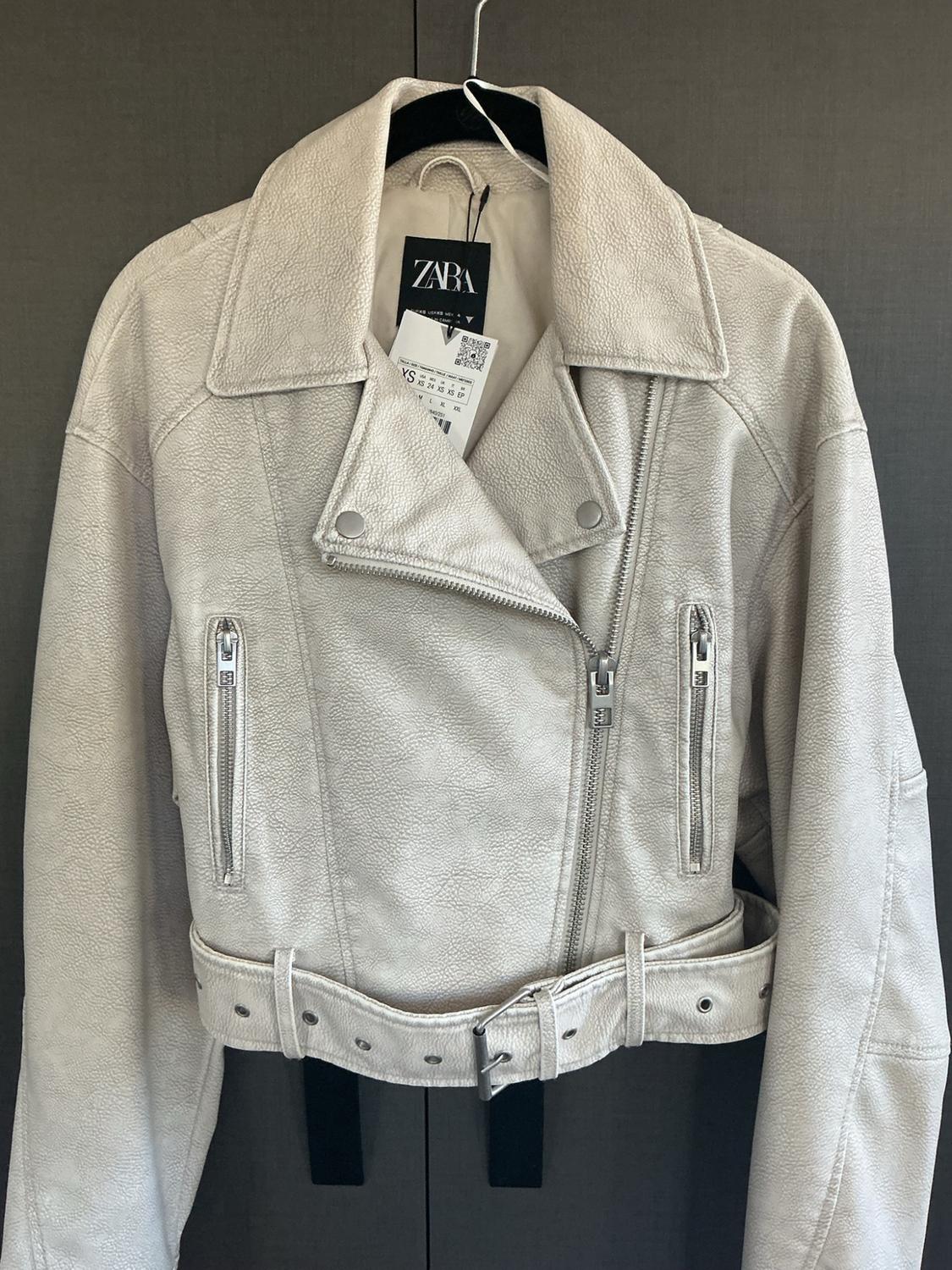 Beige jacket with zippers, belt, and ZARA label, hanging on a black hanger against a dark background.