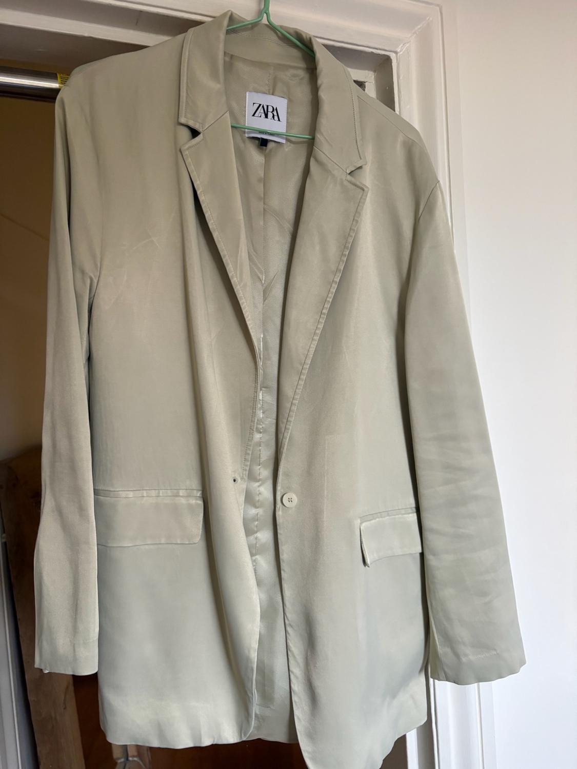 Light beige blazer hanging on a green hanger over the frame of a white door.
The label inside reads the text ZARA.