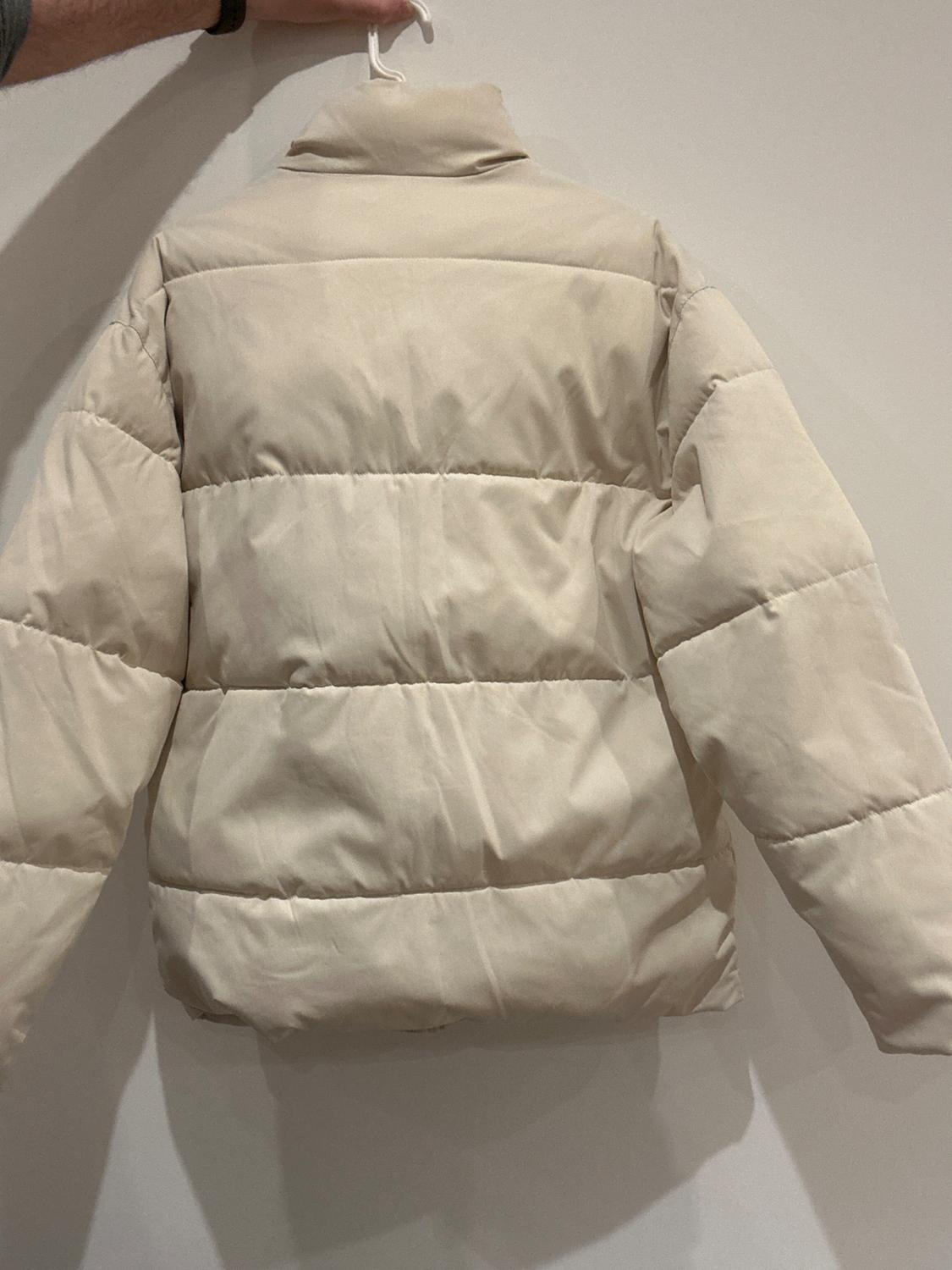 A beige padded jacket is hanging on a white hanger, seen from the back. A hand is holding the hanger in front of a white wall.