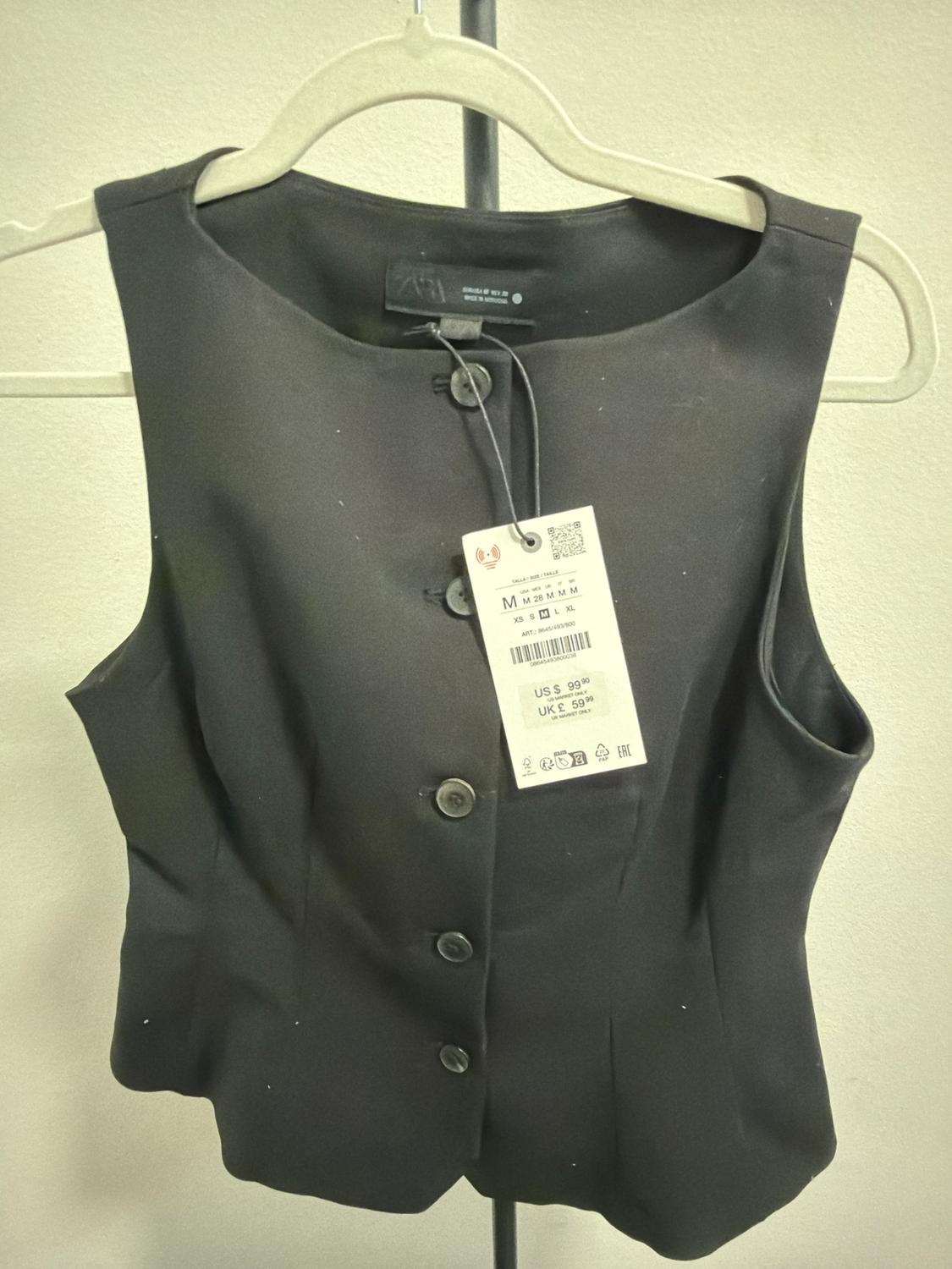Sleeveless black vest on hanger, with visible price tag: US $99.99, UK £59.99. Neutral background.