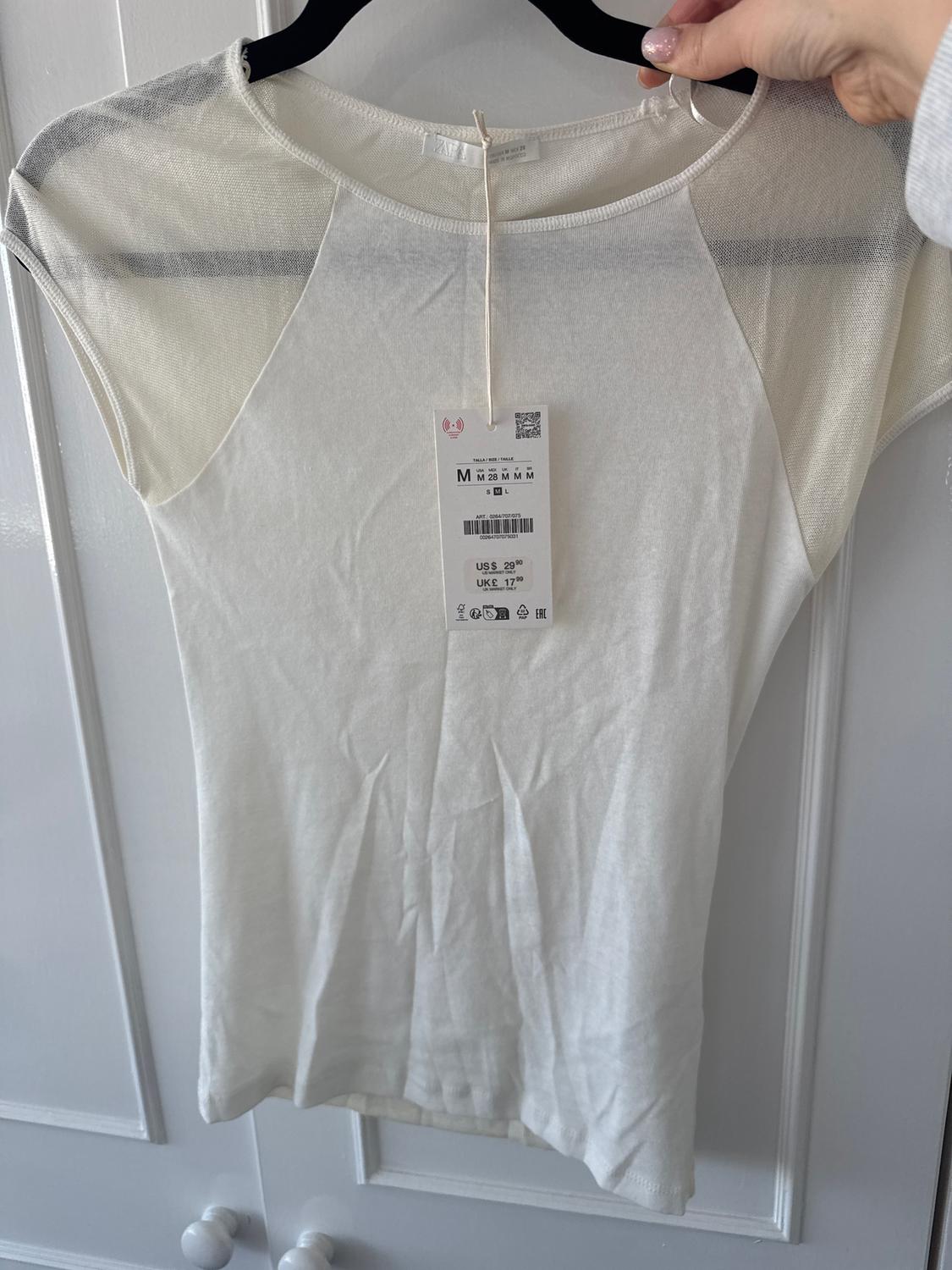 White short-sleeved t-shirt on a hanger, with a hanging tag showing size M and price US$ 29.99, UK£ 17.99. White door background.