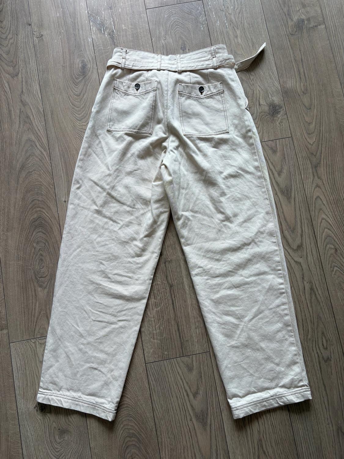 Light beige casual wide-leg trousers, with buttoned back pockets and a belt, laid out on a wooden floor.