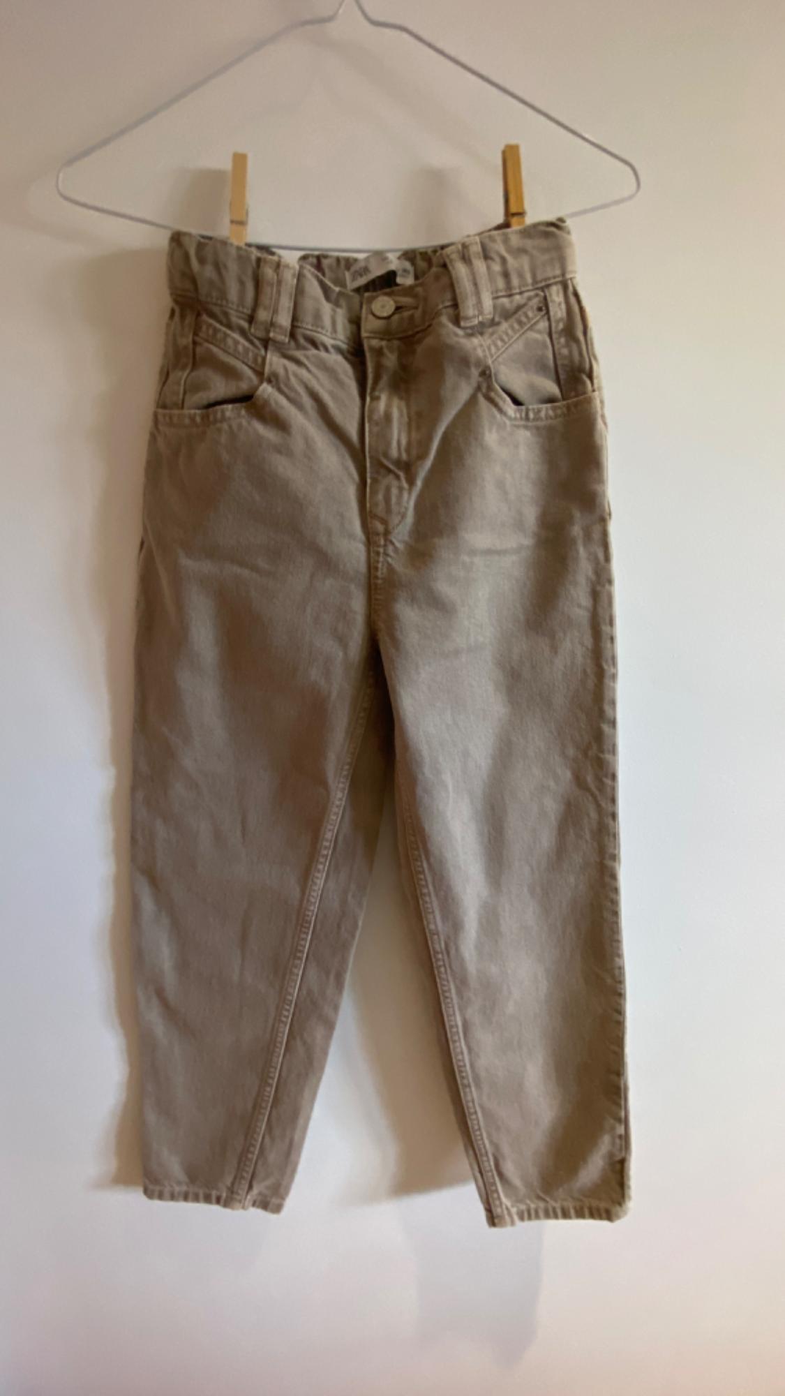Beige trousers hung on a white hanger against a white wall. Classic design with front and back pockets.