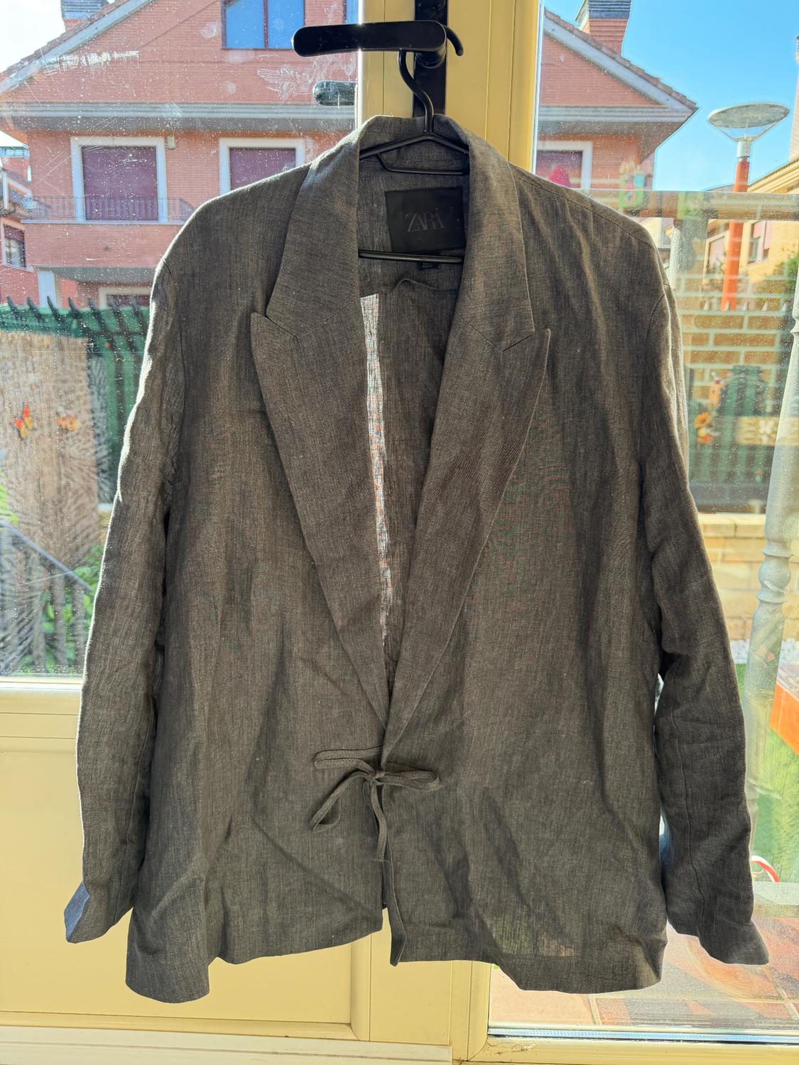 Grey linen jacket hanging on a hanger in front of a window, with a visible Zara tag. Background of houses and blue sky.