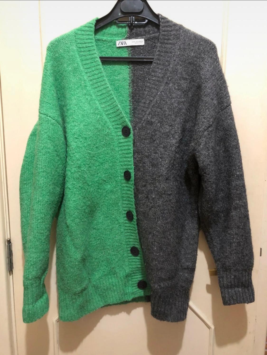 A two-tone cardigan hanging on a hanger, with one half green and the other grey, black buttons and a visible label at the neck.