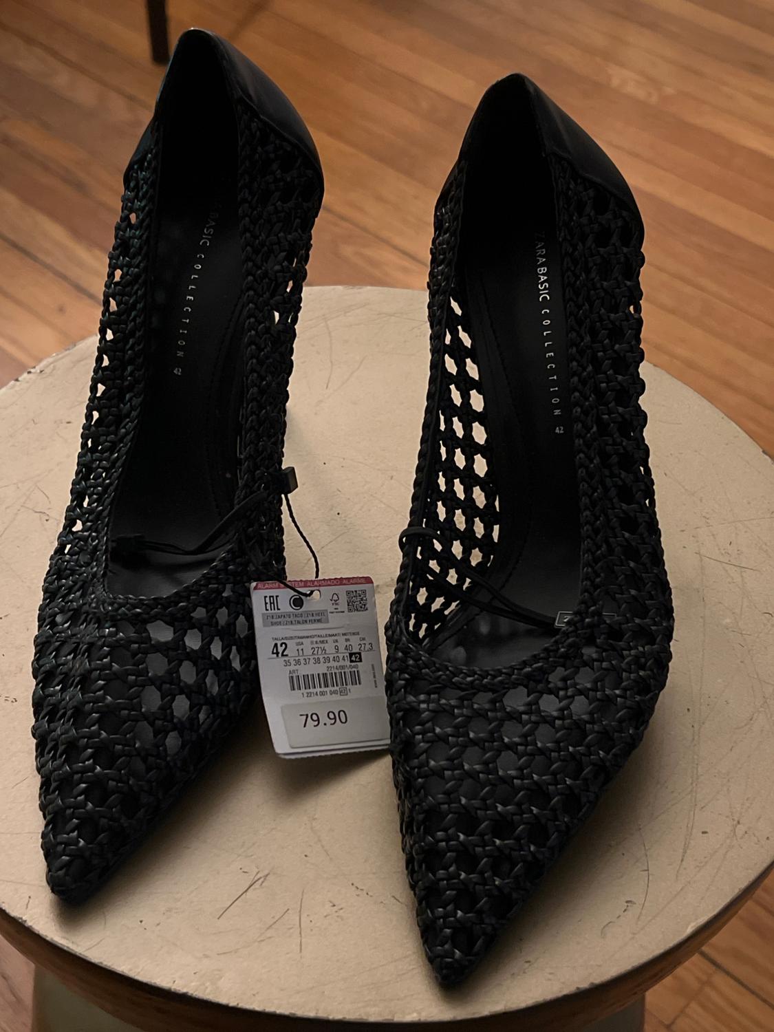Black high heels with an openwork design on a wooden table. Visible tag with price of 79.90 and size 42.