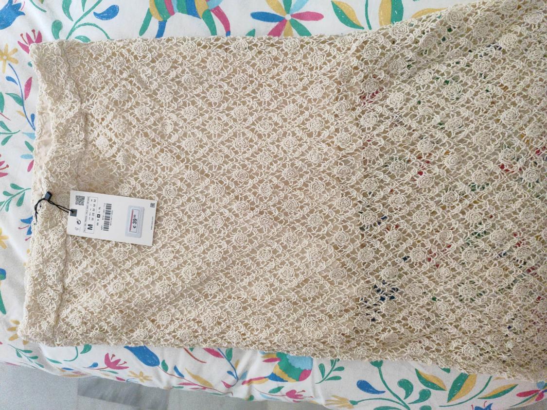 Beige lace skirt with a floral pattern on a bed with a colourful bedspread. Visible label with size M and barcode.