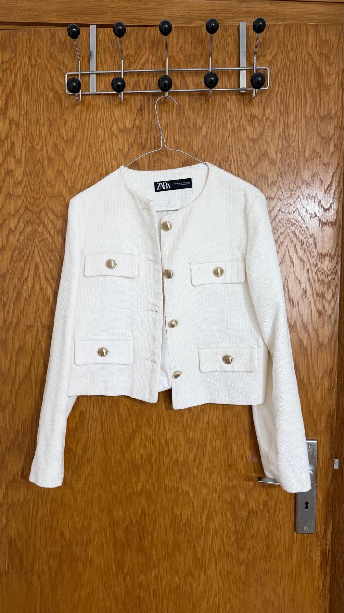 White jacket with gold buttons and four pockets, hanging on a hanger over a wooden door. Text on the label: ZARA.