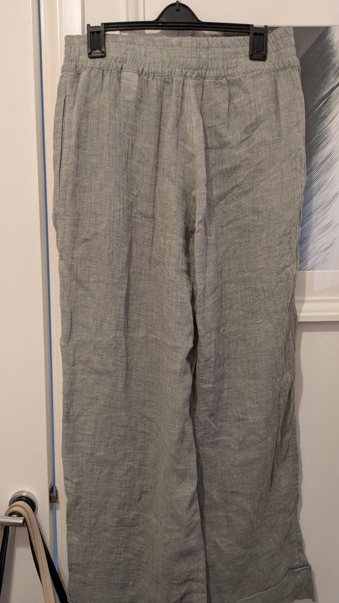 Grey linen trousers hanging on a black hanger, with an elasticated waist, against a white wall background and a partially visible picture.