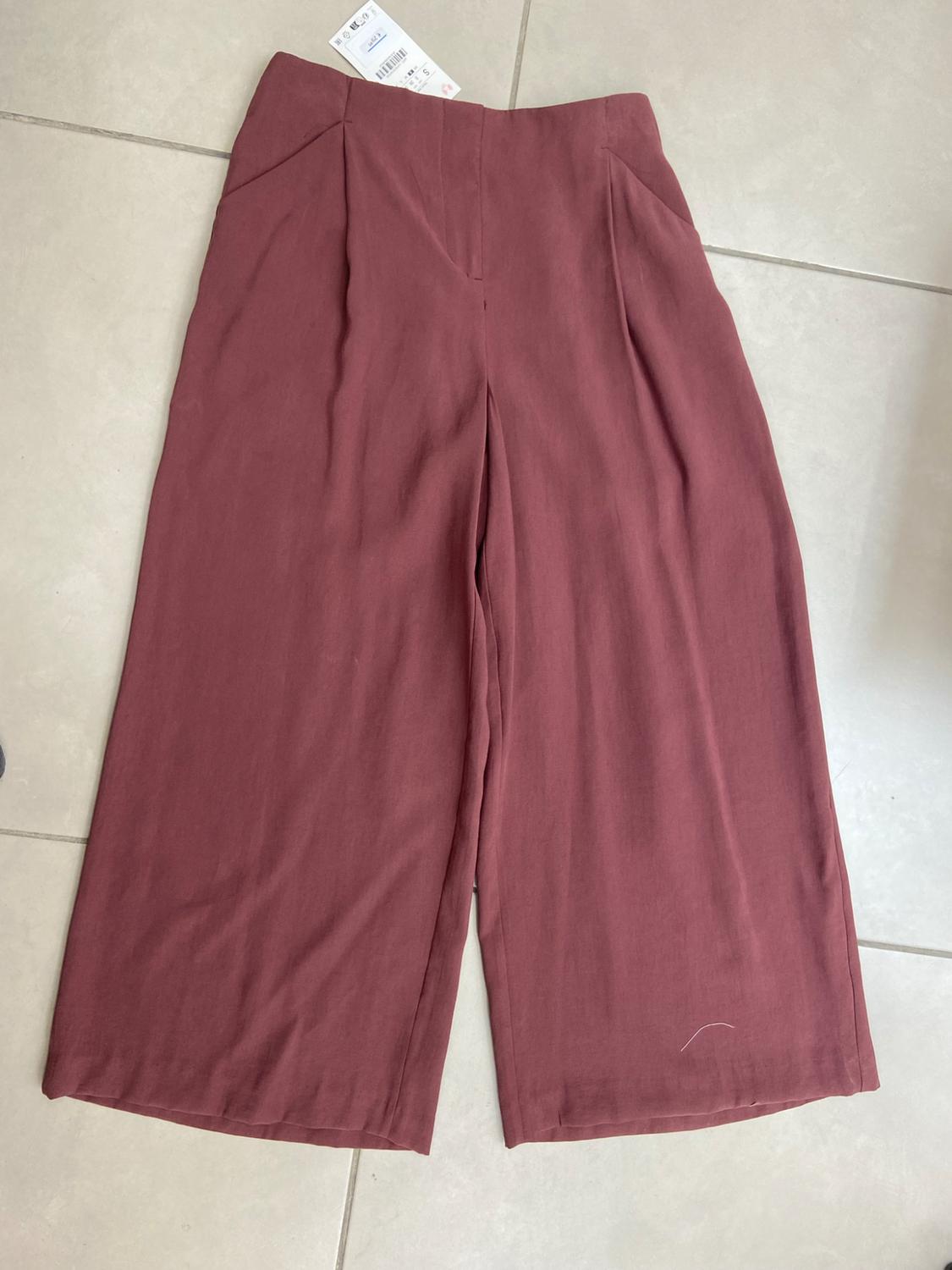 High-waisted, wide-leg burgundy trousers with pleats and pockets, laid out on a light-tiled floor with a label at the waistband.