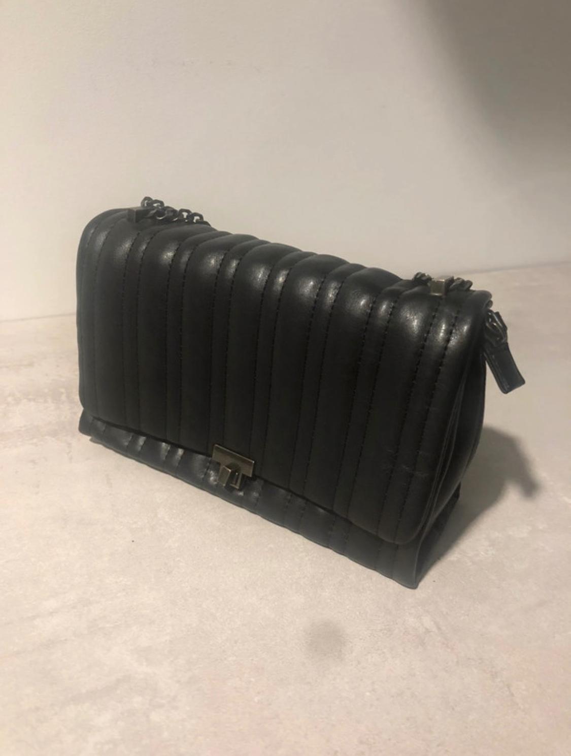Black quilted bag with vertical stitching, a metal front fastening and a chain handle, resting on a light-coloured surface against a neutral background.