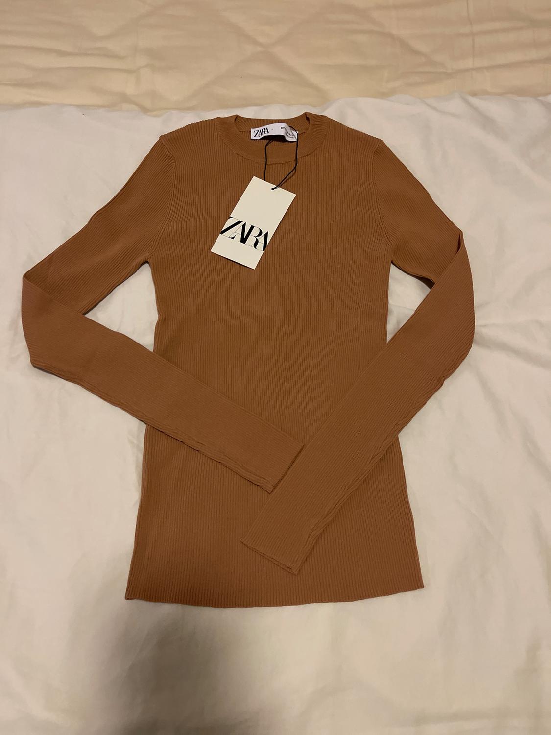 Brown long-sleeved jumper on a white bed, with a hanging tag that says "ZARA".