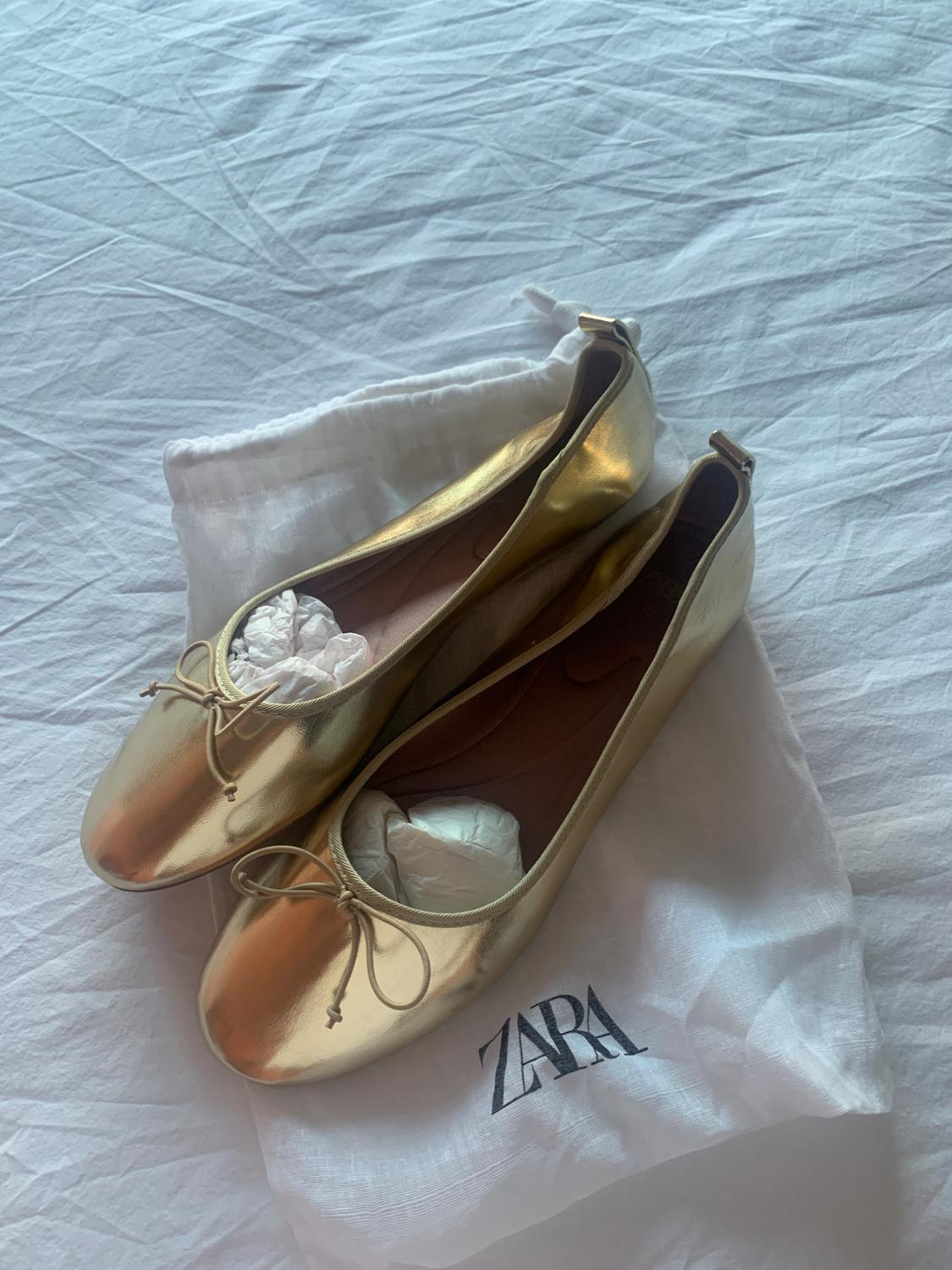 Gold ballet flats with a bow on a white fabric bag with the text ZARA, resting on a rumpled white sheet.