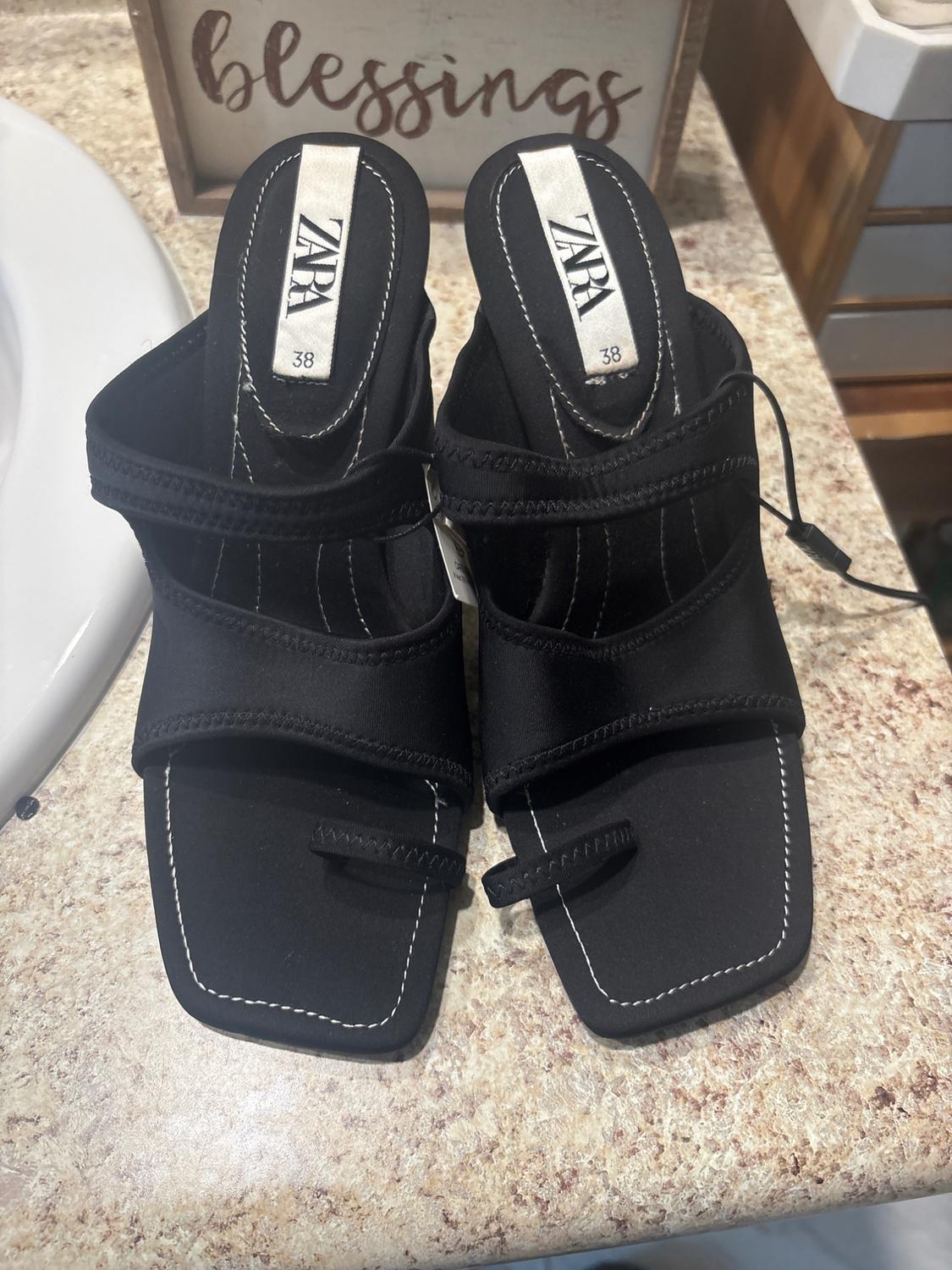 Black square-heeled sandals with white stitching and a "ZARA 38" tag, on a marble surface. "Blessings" sign in the background.