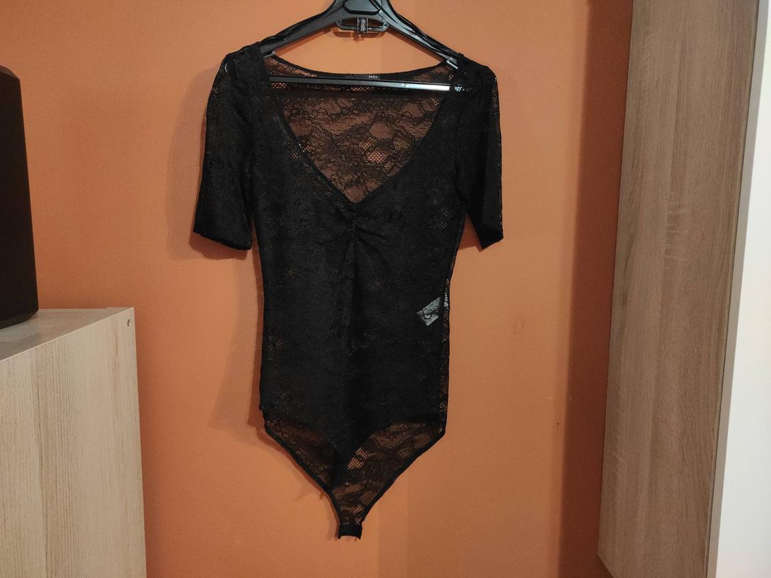 Black lace bodysuit hanging on a hanger against an orange wall, with short sleeves and a V-neck.