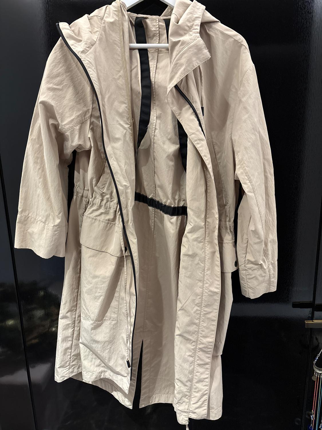 Beige coat with hood and black drawstring hanging on a hanger, dark background.