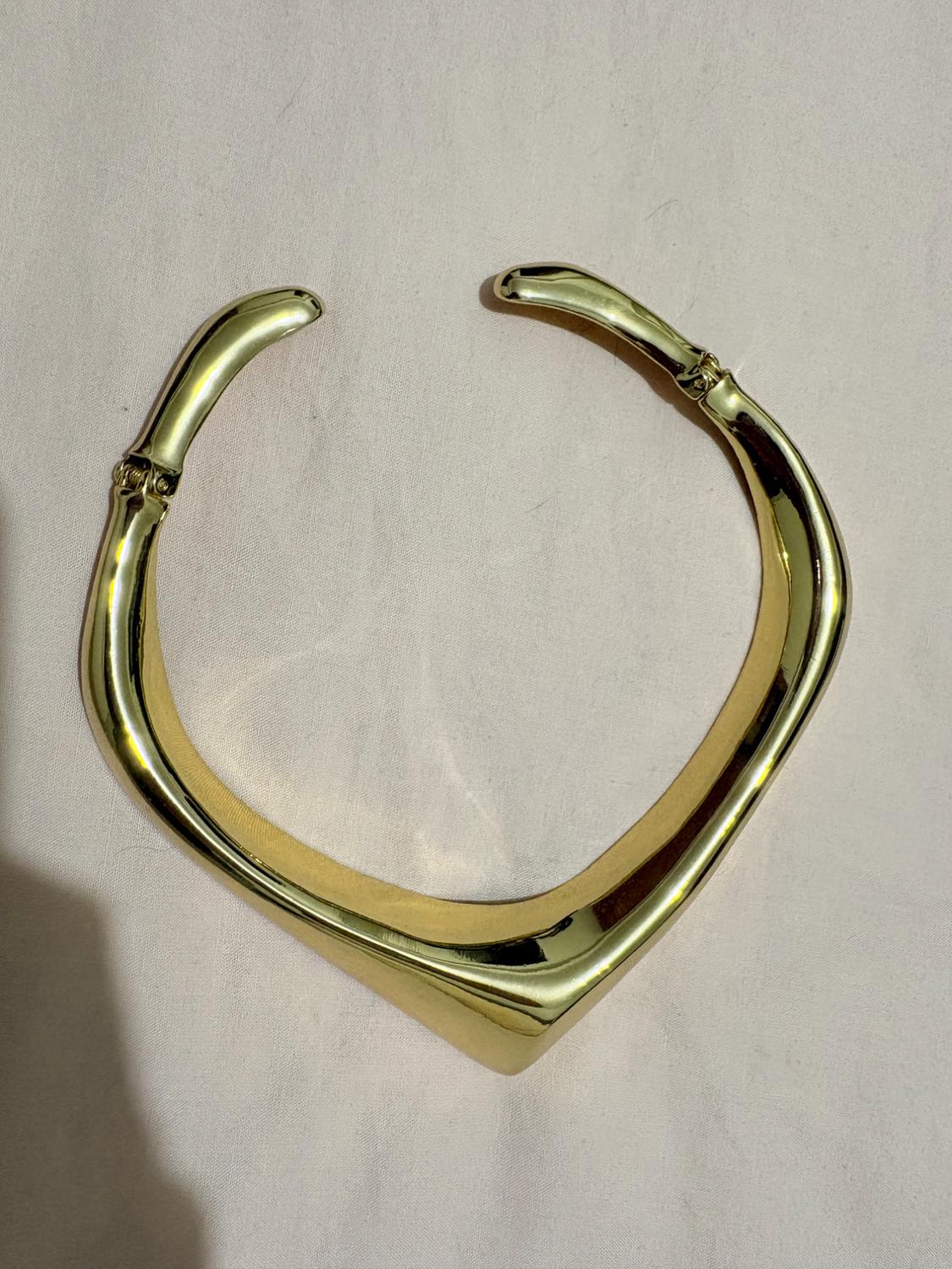 Gold-tone rigid collar necklace with a pointed front design and side hinges, resting on a light pink fabric background.