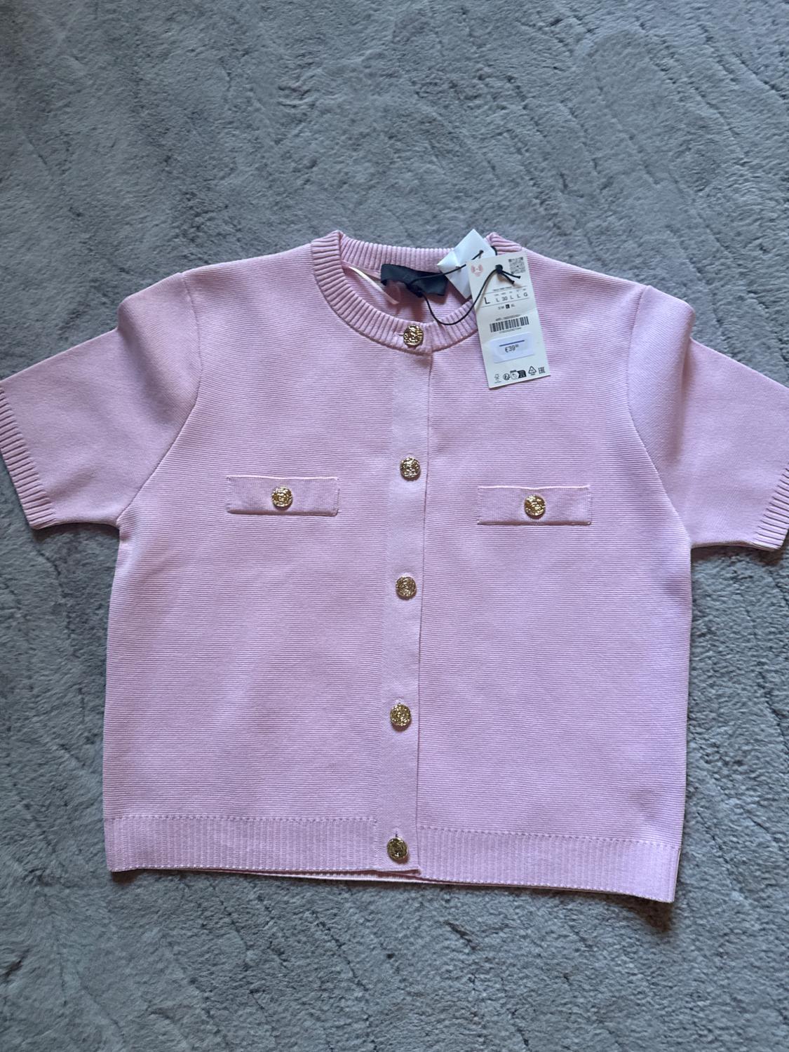 Light pink, short-sleeved jacket with gold buttons and two front pockets. Tags hanging from the collar. Grey background.