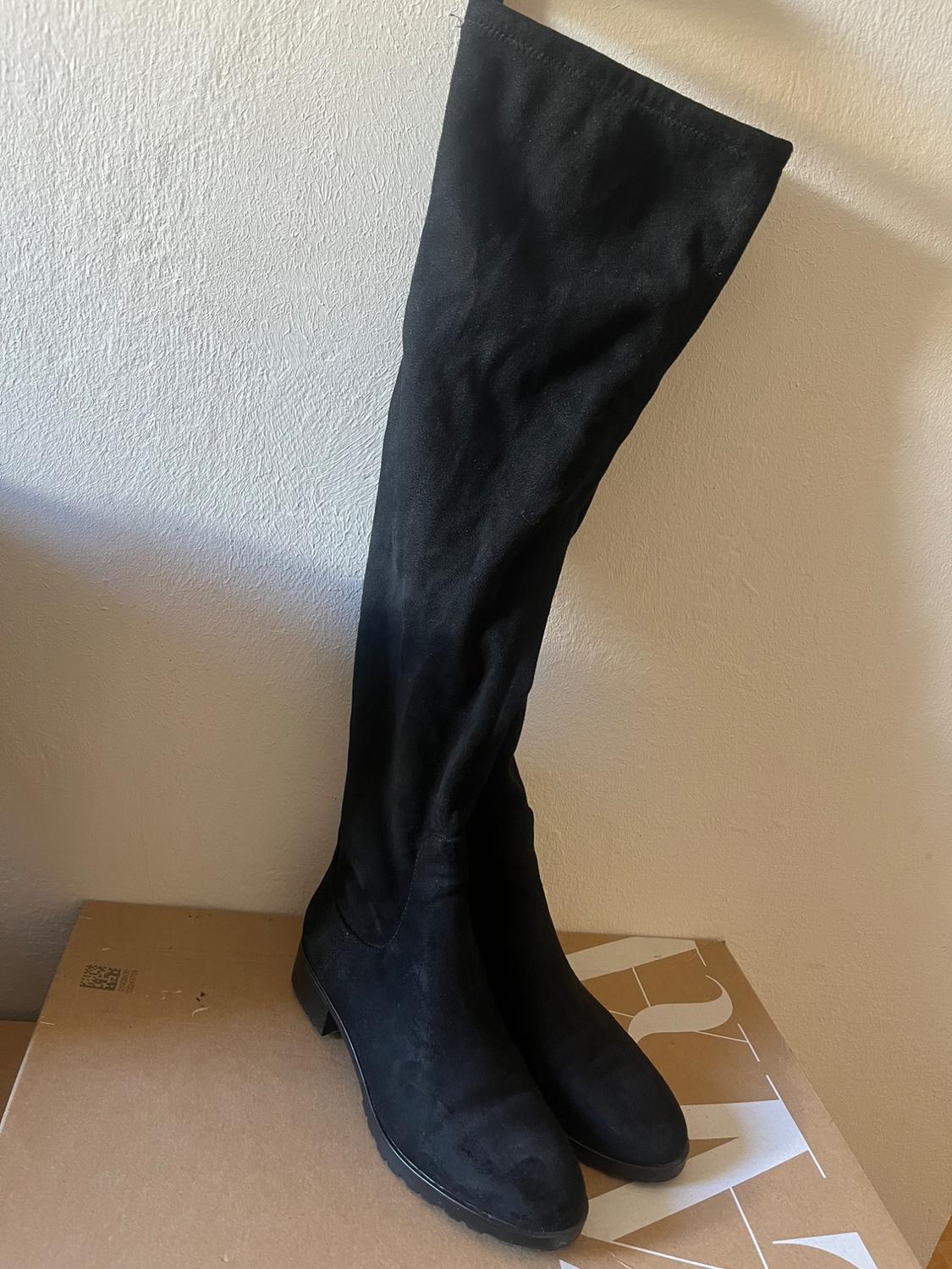 Black suede knee-high boots on a cardboard box, leaning against a white wall.