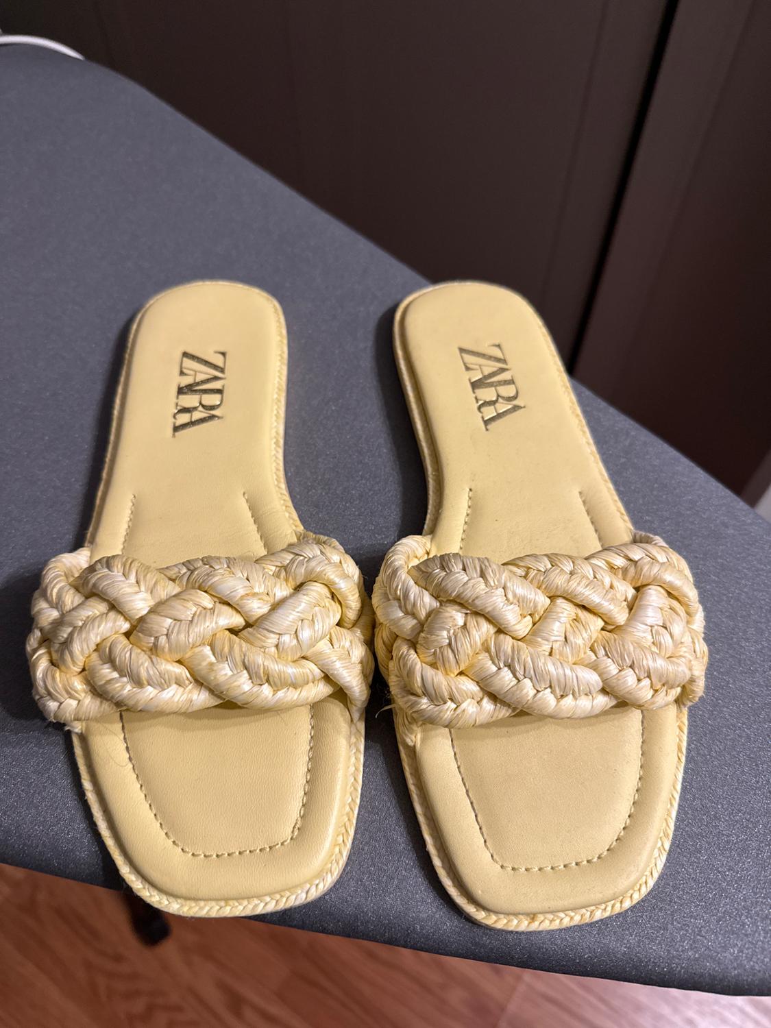 Yellow flat sandals with thick braided straps on top. The insole features the "ZARA" logo. Gray and brown background.