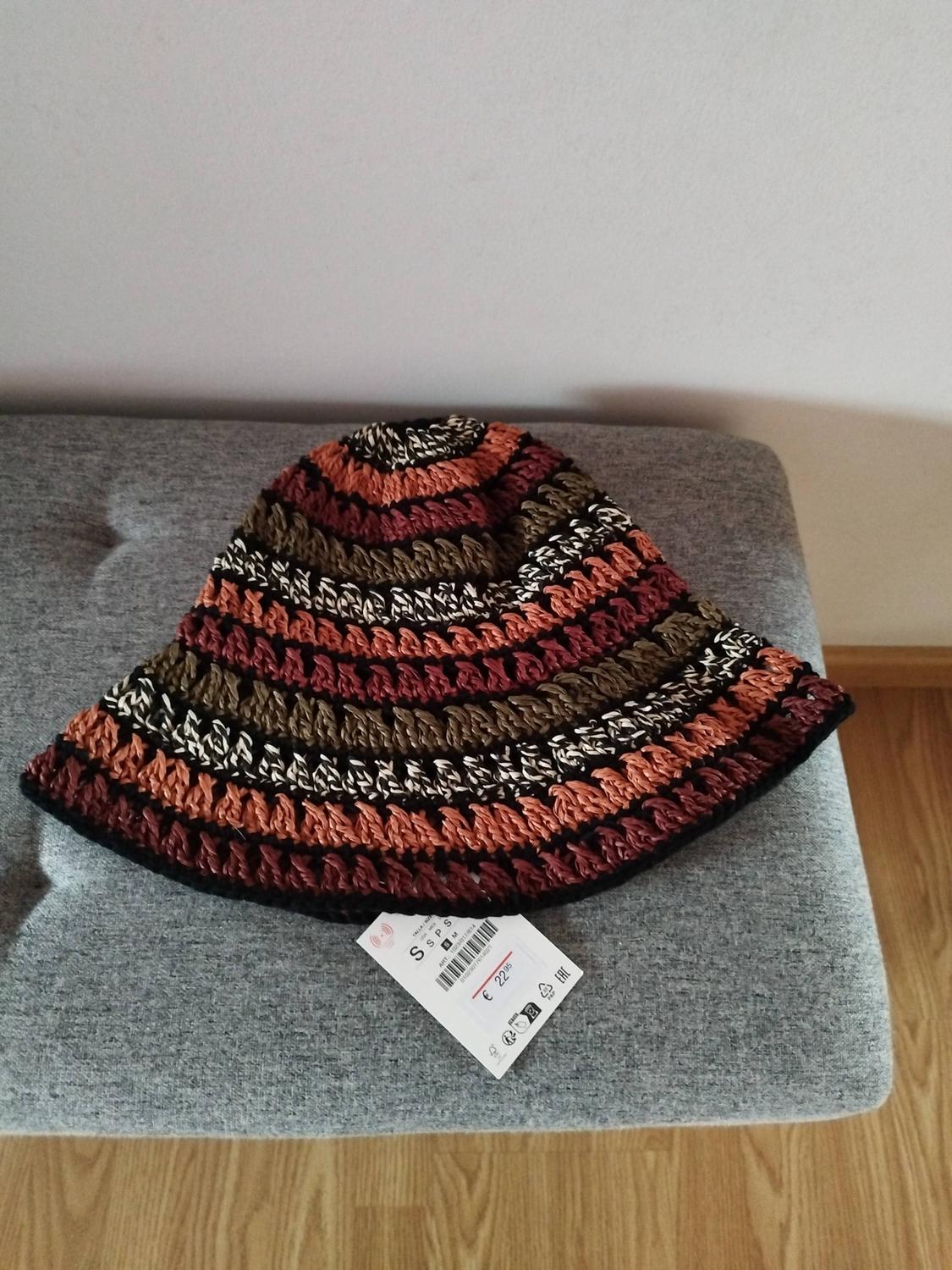 Crochet hat with brown, orange, and black stripes on a grey bench. Visible tag with a price of €22.