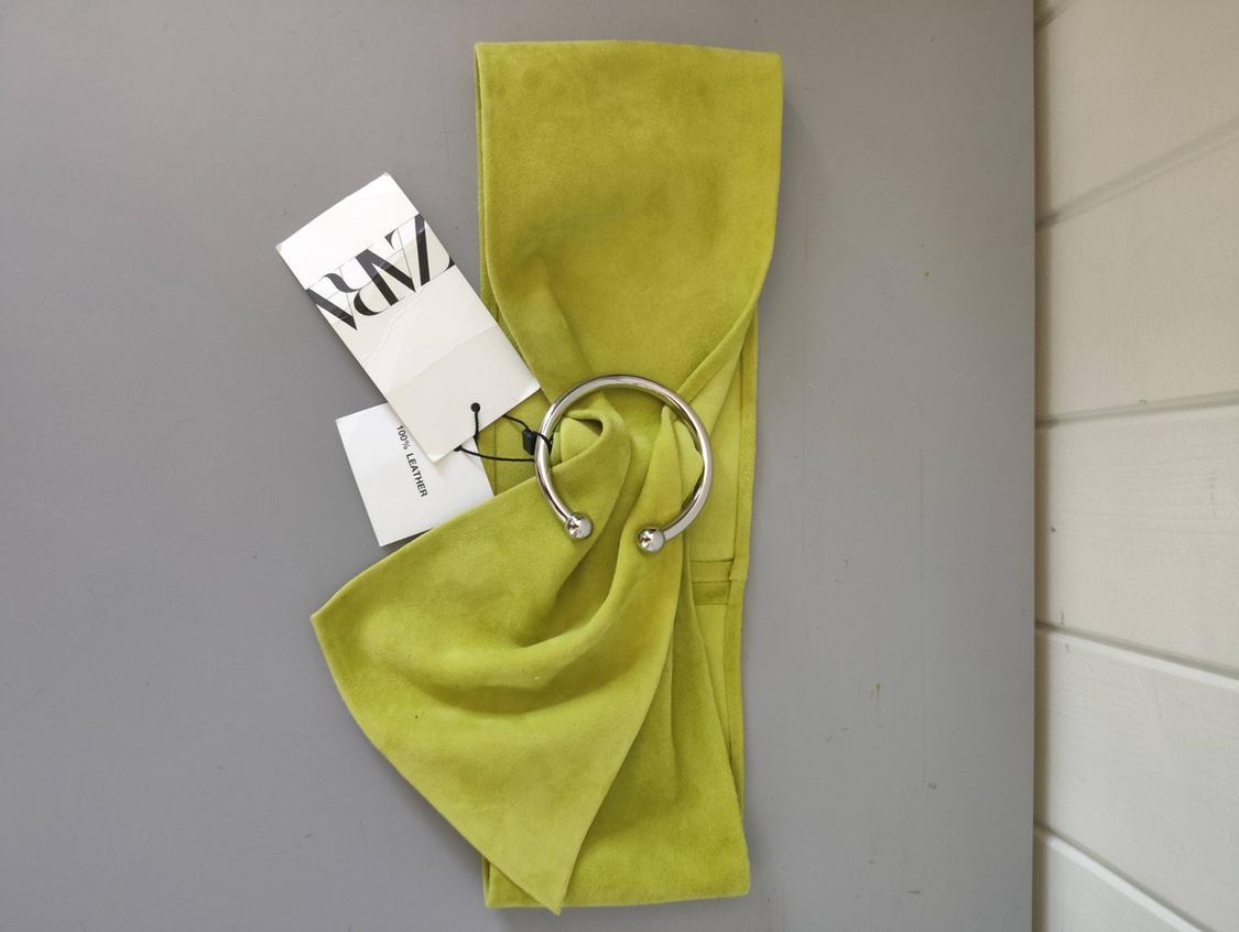 Green belt with a silver-coloured metal ring on a grey background. Includes labels with the text ZARA and 100% LEATHER.
