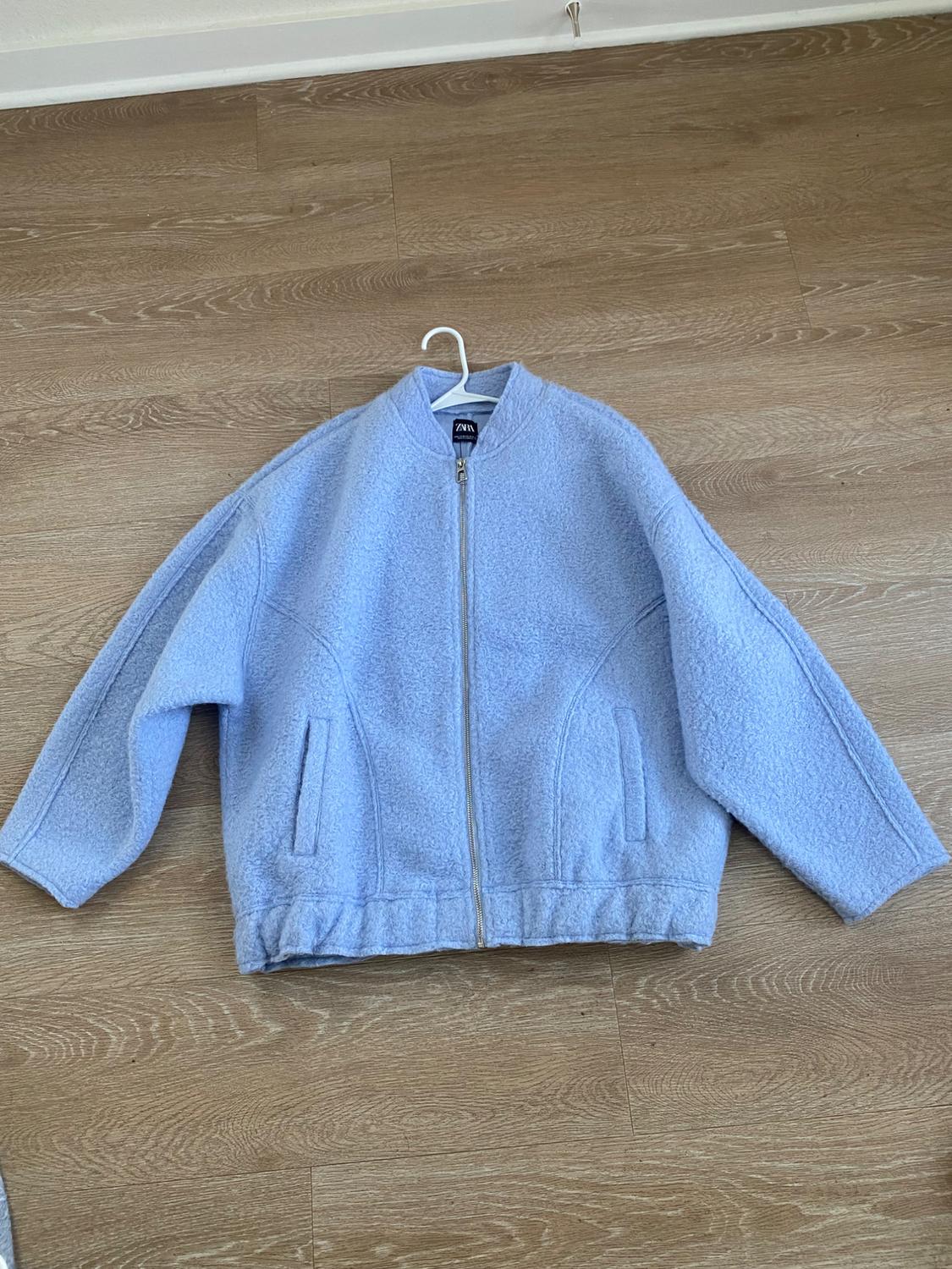 Light blue jacket with a zipper and front pockets, hanging on a hanger over a light wood floor. Visible tag.