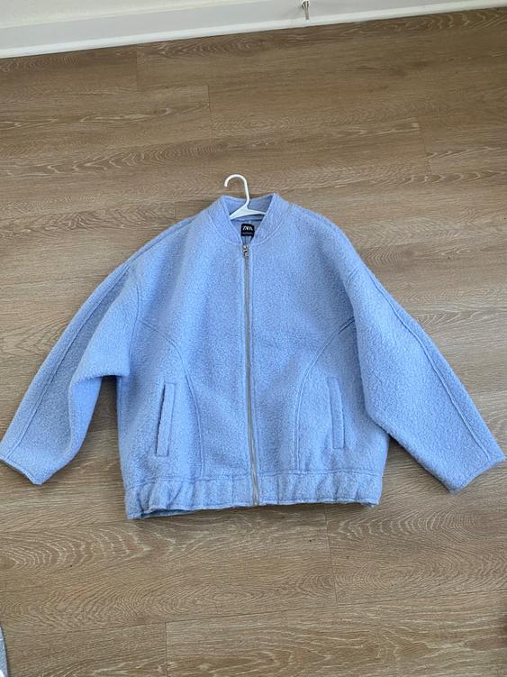 Light blue jacket with a zipper and front pockets, hanging on a hanger over a light wood floor. Visible tag.