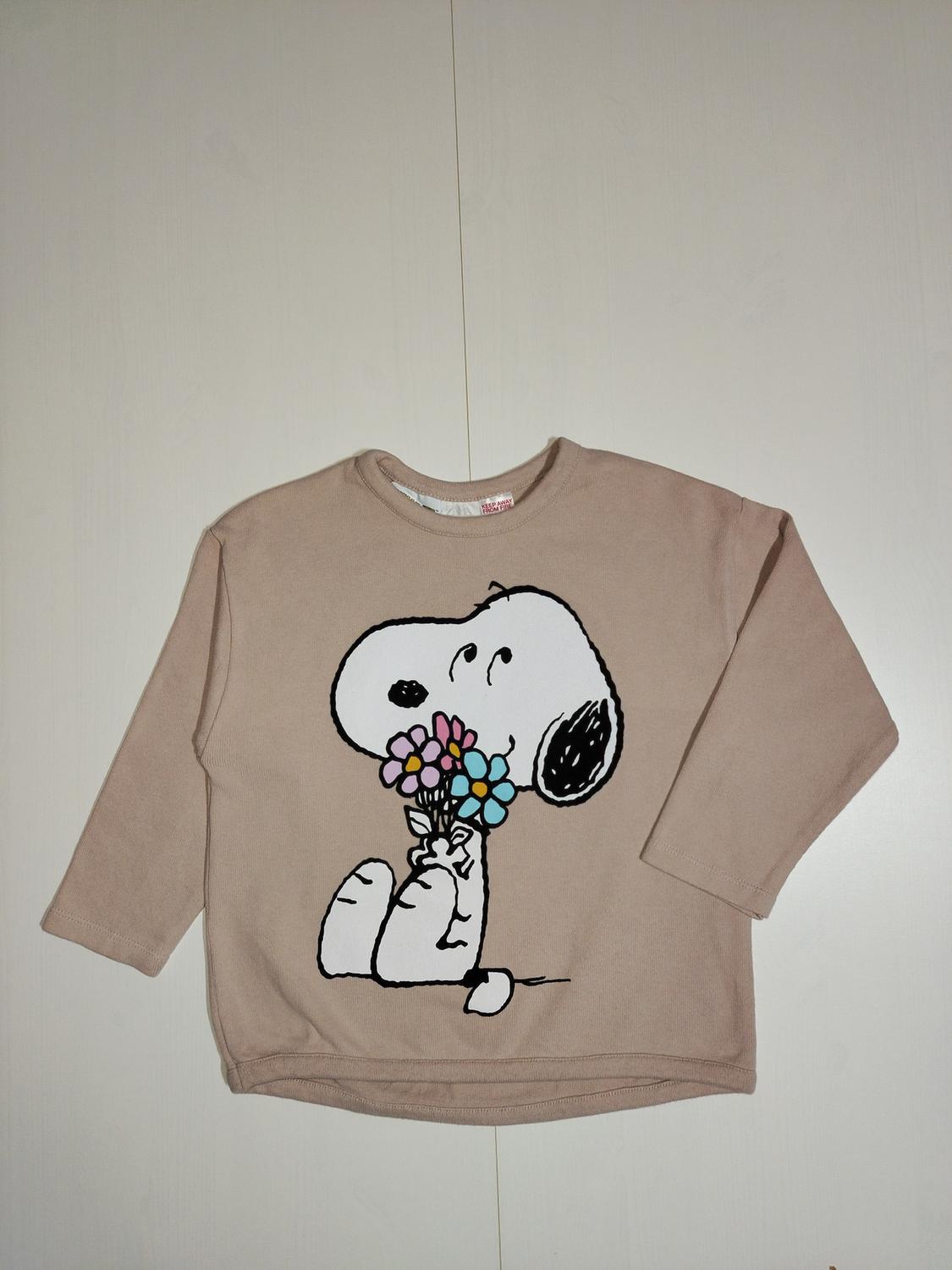 Beige sweatshirt with a cartoon character design holding colourful flowers. White background.