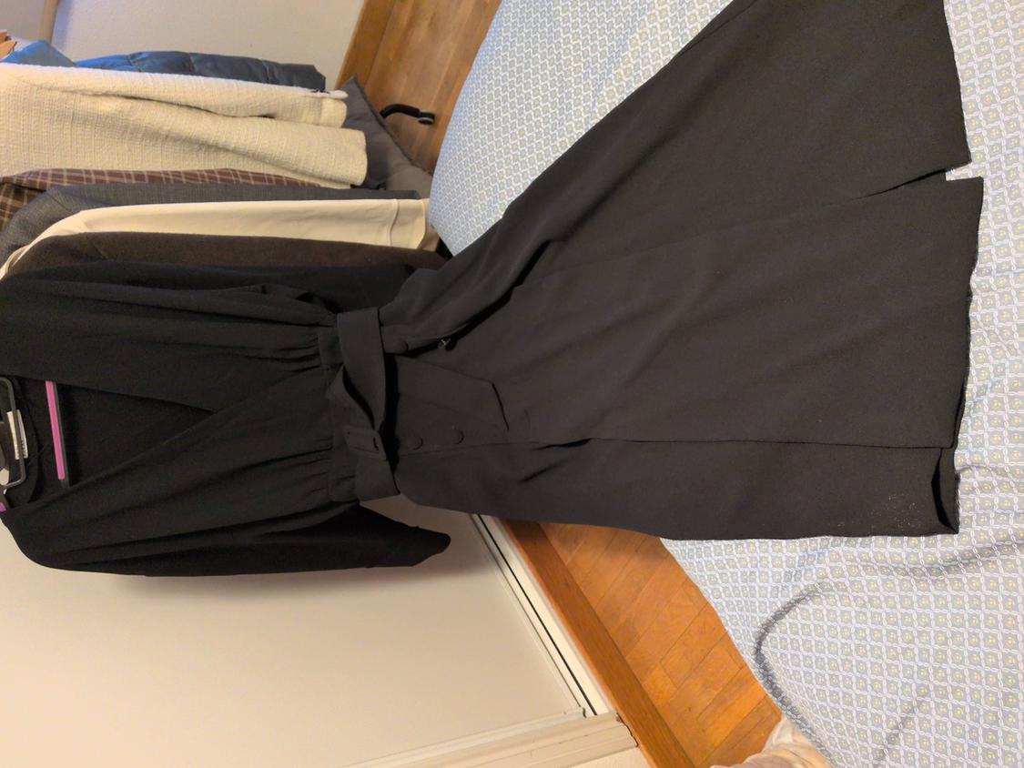 Black dress with belt on a bed, next to several garments hanging on hangers. White wall and wooden floor in the background.