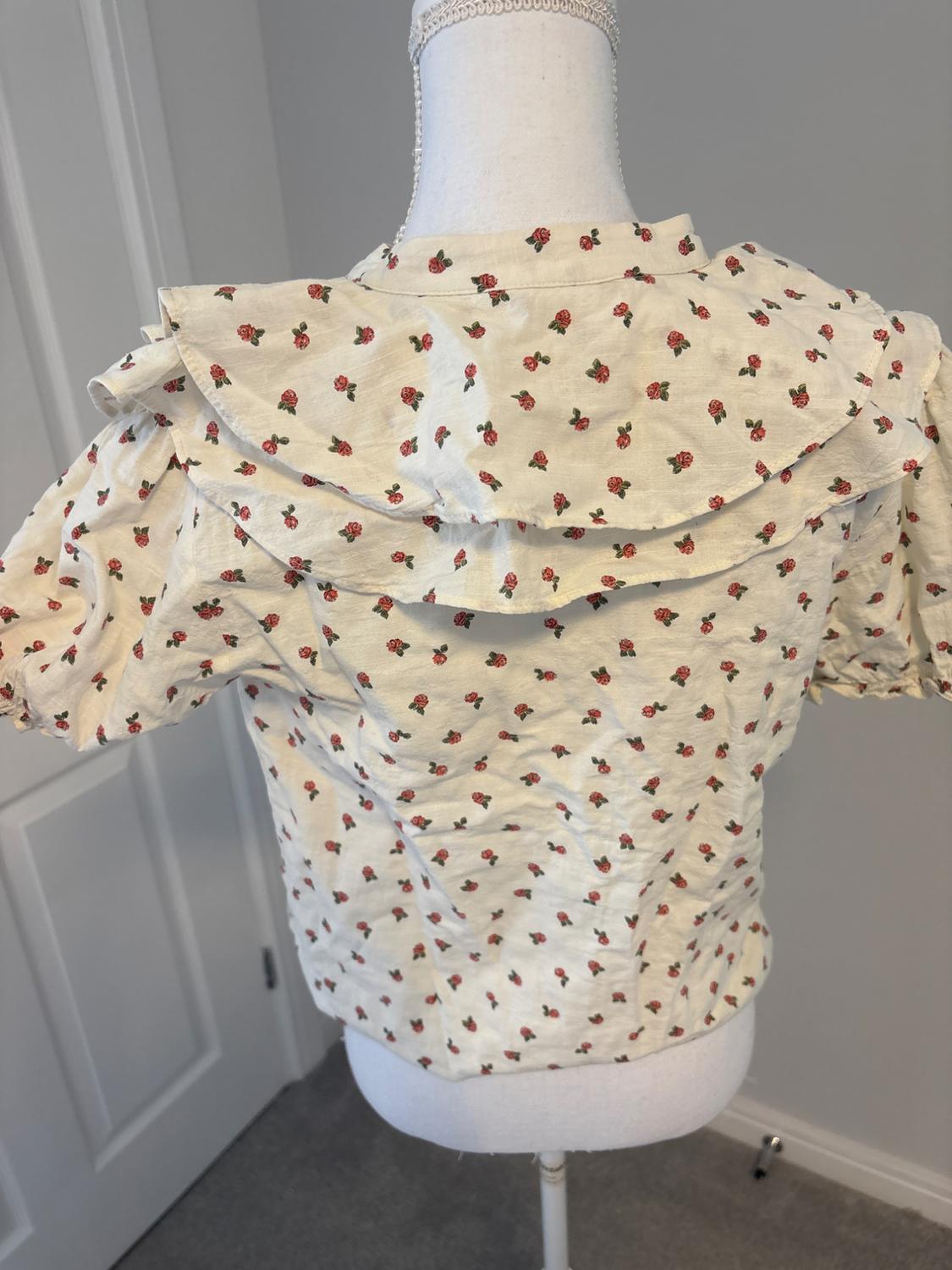 Rear view of a cream blouse with a red rose print, short sleeves and a frilled collar on a mannequin in a grey room.