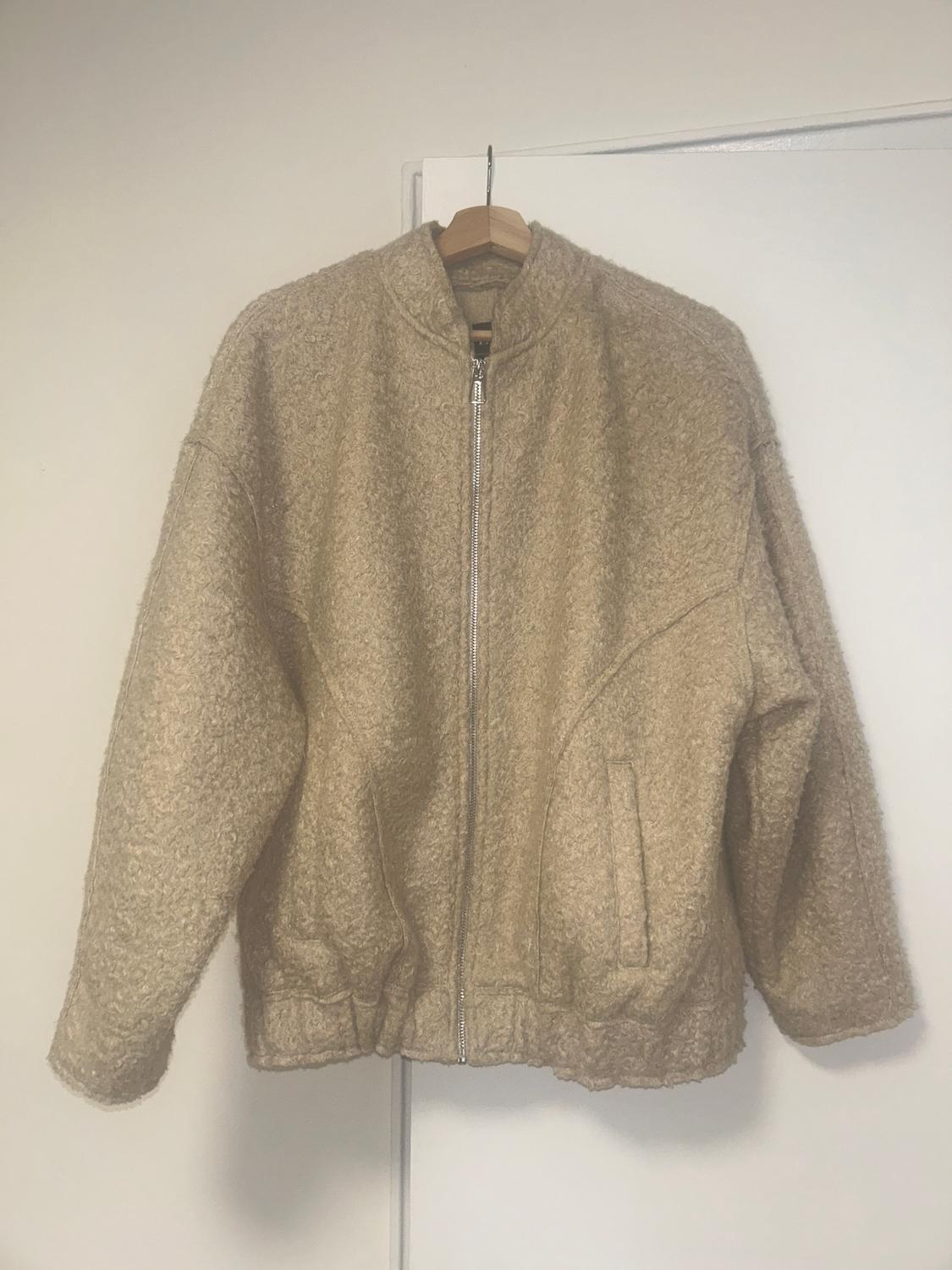 Soft-textured beige jacket hanging on a wooden hanger, with a front zip and side pockets. White background.