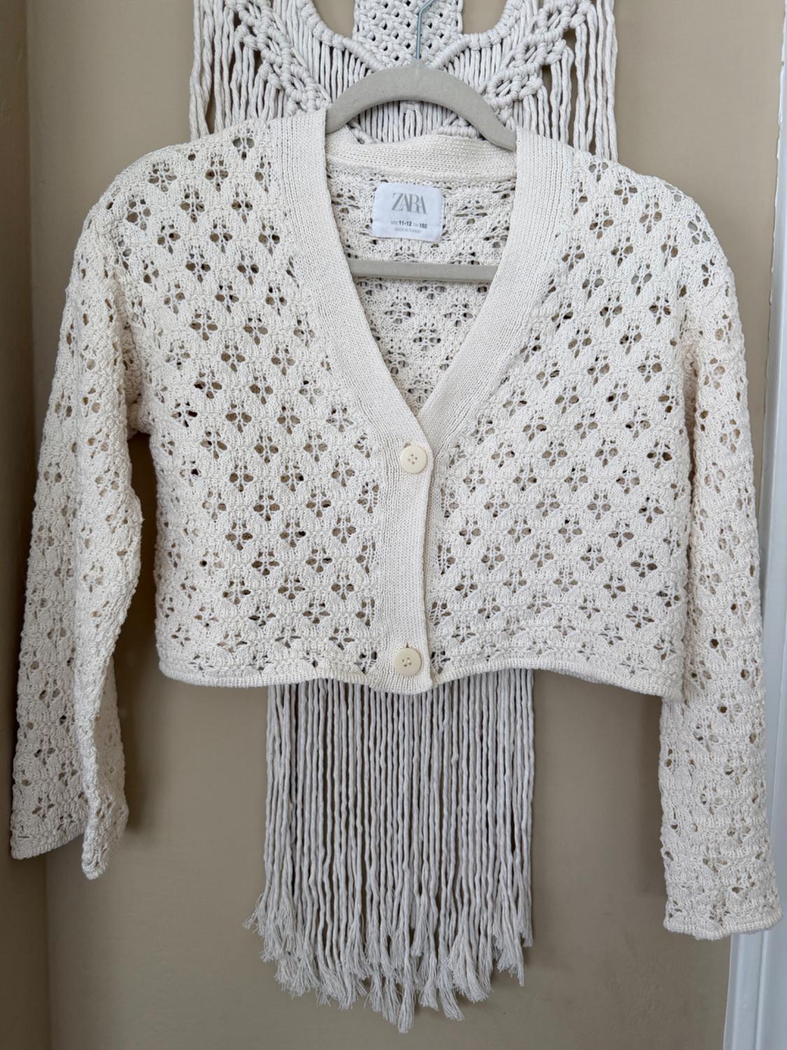 White crochet cardigan with buttons, hanging on a hanger against a macrame background. Visible tag: ZARA.