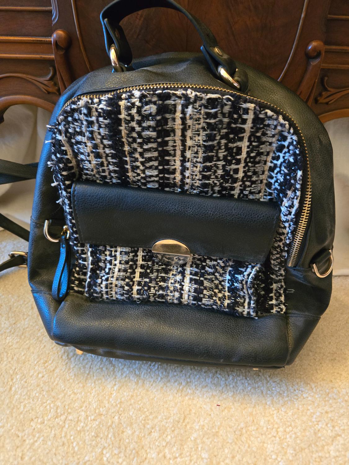 Black backpack with black and white woven details, gold zip, and front pocket, on a beige rug with a wooden cabinet in the background.
