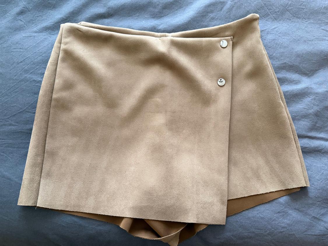 Short beige skirt with a wrap-around design and two decorative buttons at the top, on a light grey background.