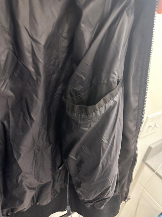 Detail of a black jacket with an open zipper, showing the interior and a pocket. The fabric has a wrinkled finish.