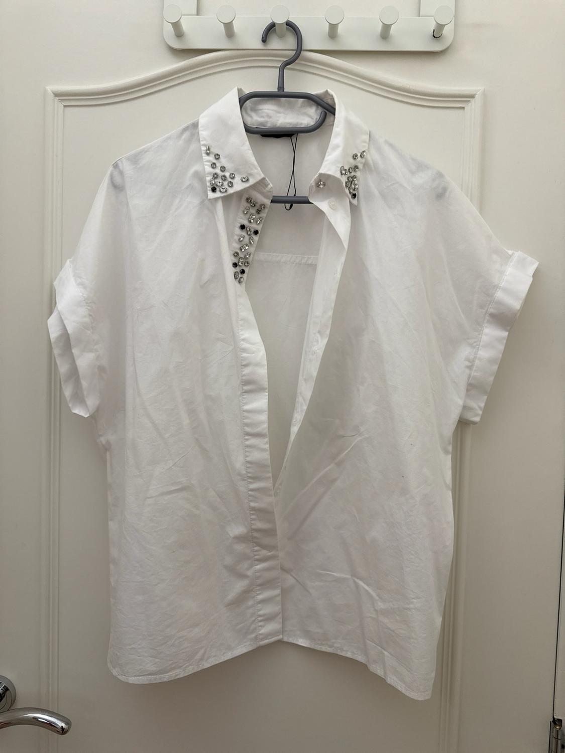 Unbuttoned short-sleeved white shirt with beaded details on the collar, hanging on a hanger over a white door.