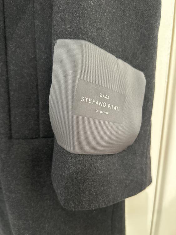 Detail of a dark grey coat with a label that reads "ZARA STEFANO PILATI COLLECTION" on a light grey patch.