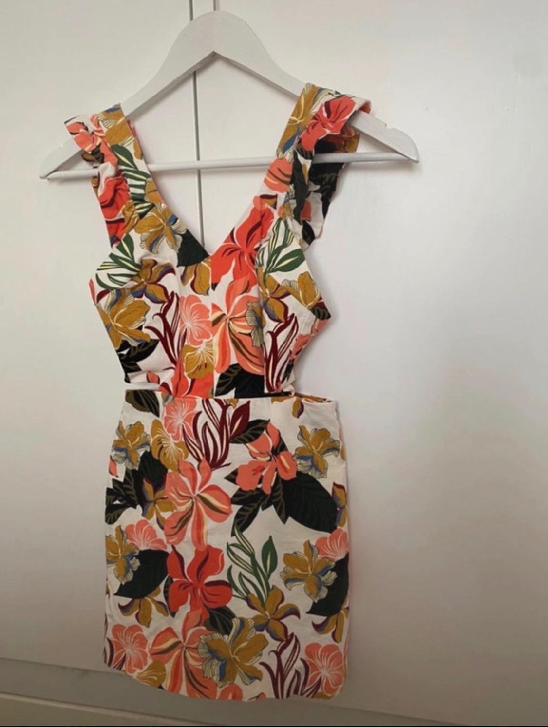 Sleeveless dress with a floral print in orange, yellow, and green tones, hanging on a white hanger in front of a white wall.
