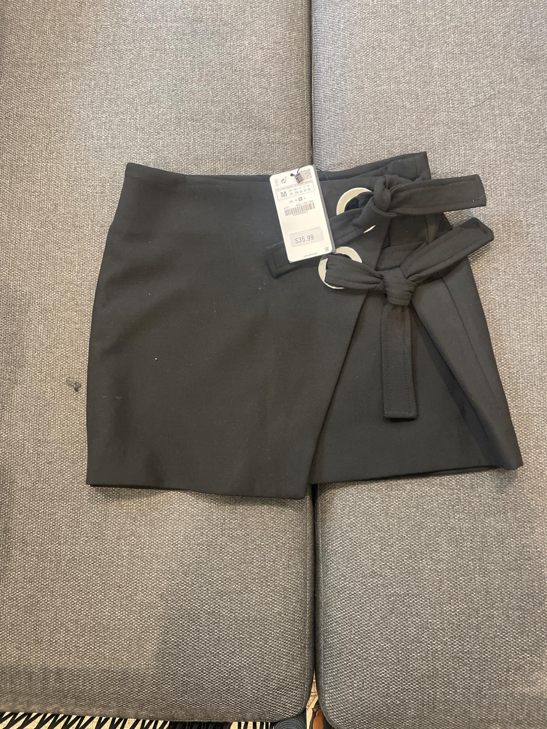 Black skirt with bow and metallic eyelets on grey sofa. Visible tag with price of $35.99.