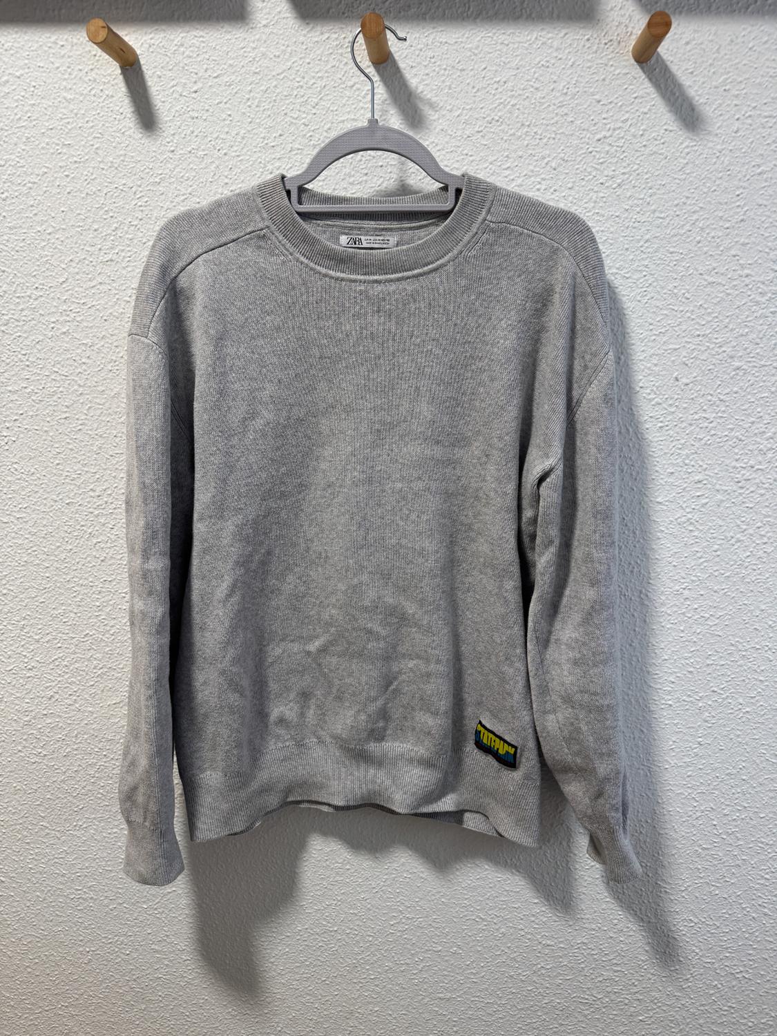 Grey jumper hanging on a hanger against a white wall. Label visible in the bottom right corner.