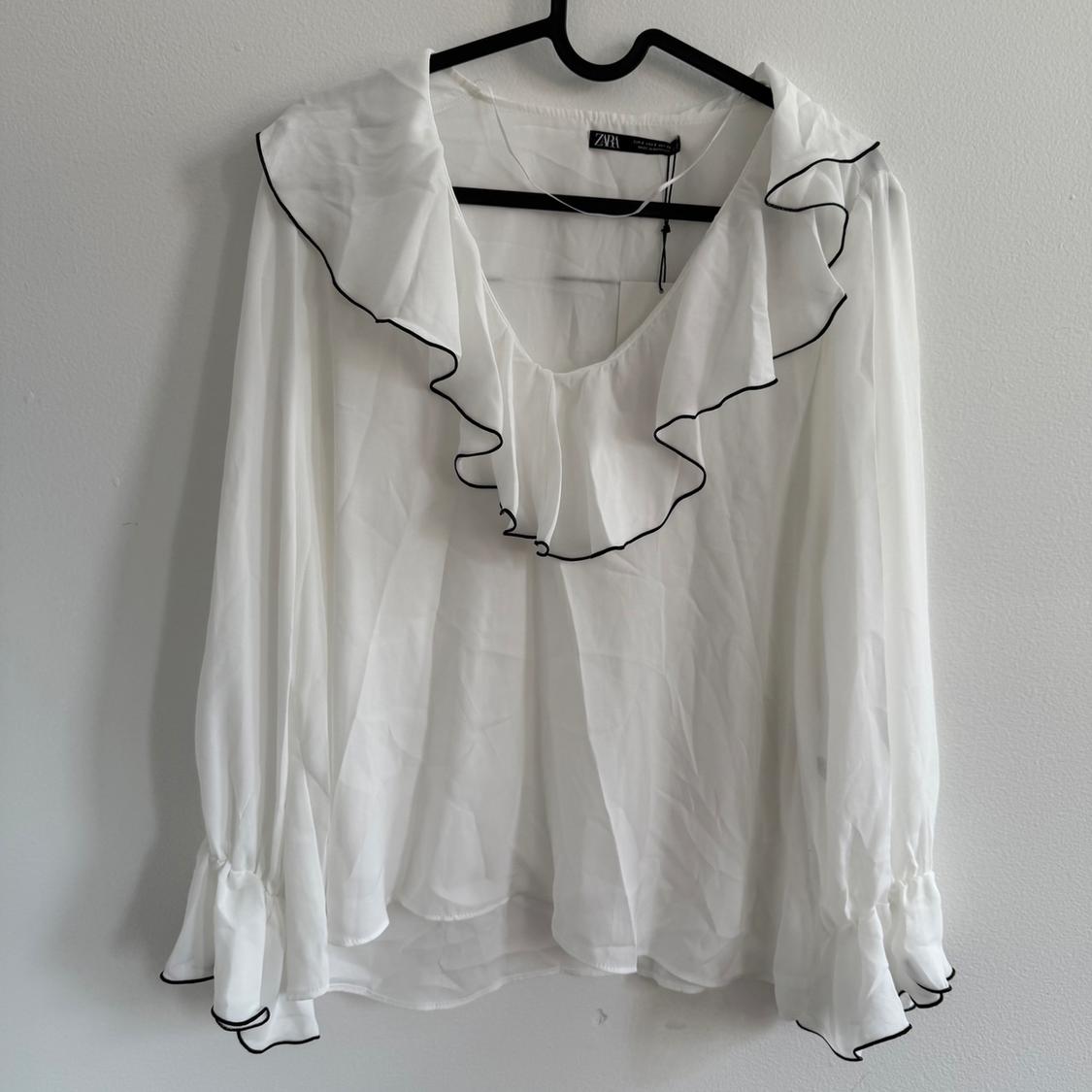 White long-sleeved blouse with ruffles at the neck and cuffs, black trim details, hanging on a hanger against a white wall.
