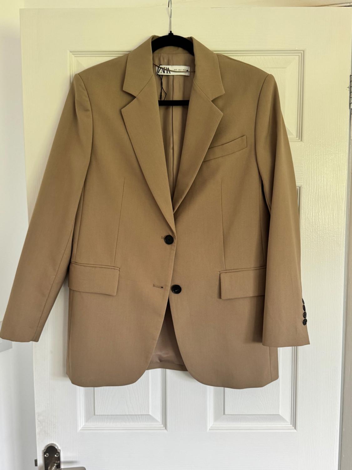Beige jacket hanging on a white door, with classic lapels and two black buttons. Label visible on the inside of the collar.
