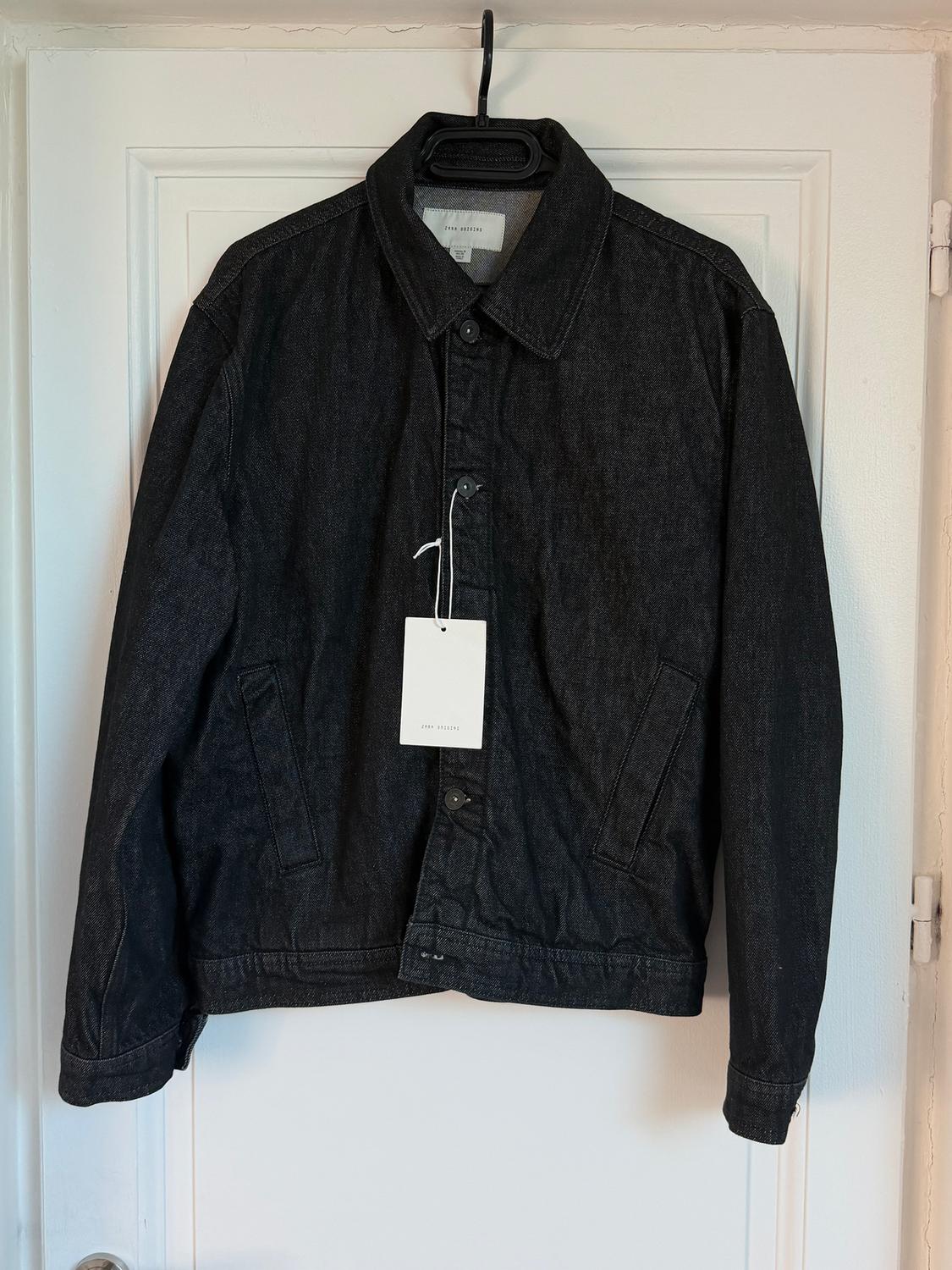 Black denim jacket hanging on a white door, with a white label that reads "Zara Origins".