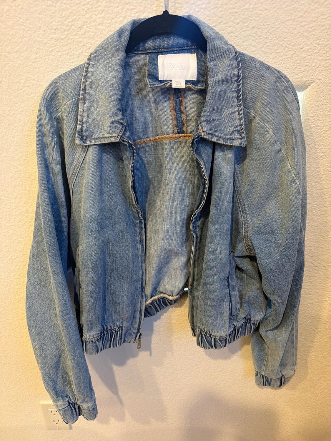 Light blue denim jacket with a zipper, elastic collar, and hem. Hanging on a black hanger against a white wall. ZARA tag visible.