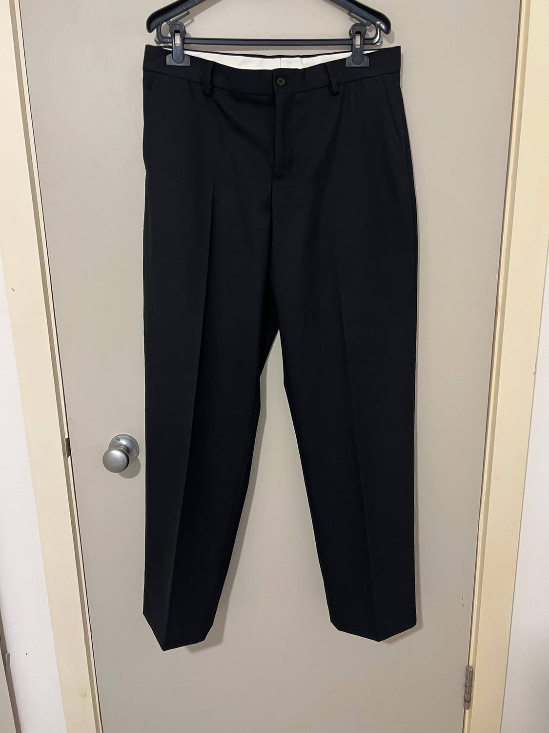 Black trousers hanging on a hanger over a beige door. Classic design with front pleats and button fastening.
