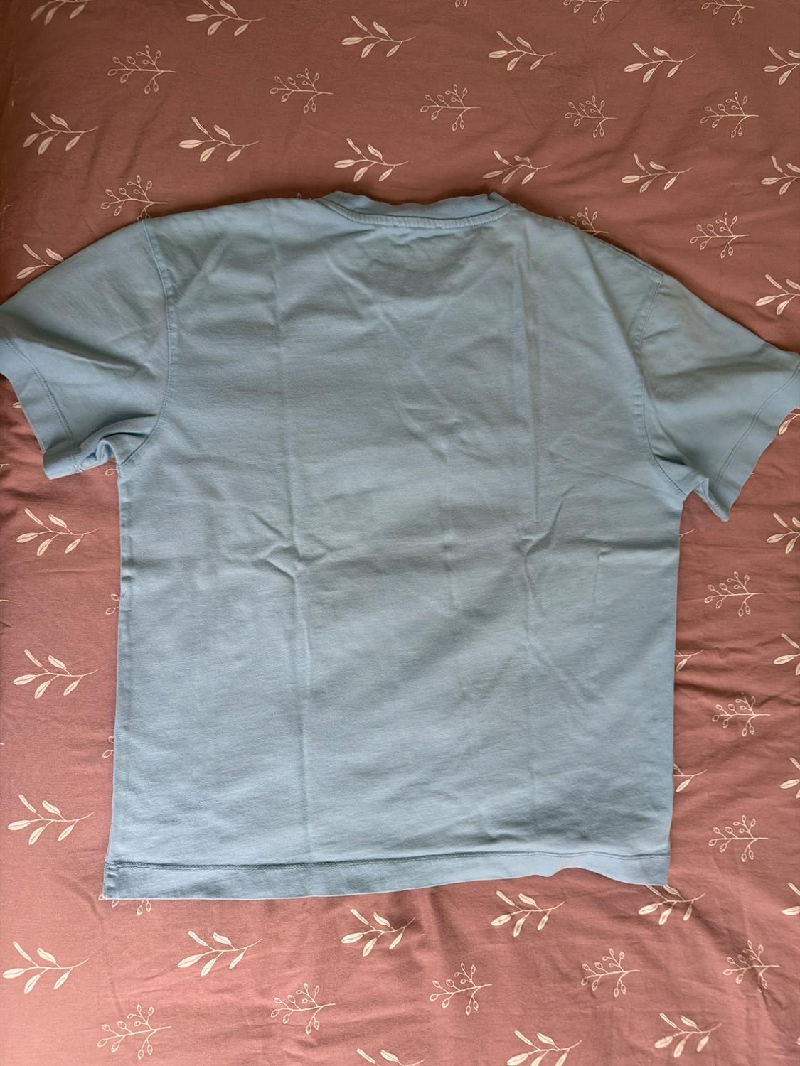 Light blue short-sleeved T-shirt seen from behind, laid out on a pink fabric surface with a white leaf print.