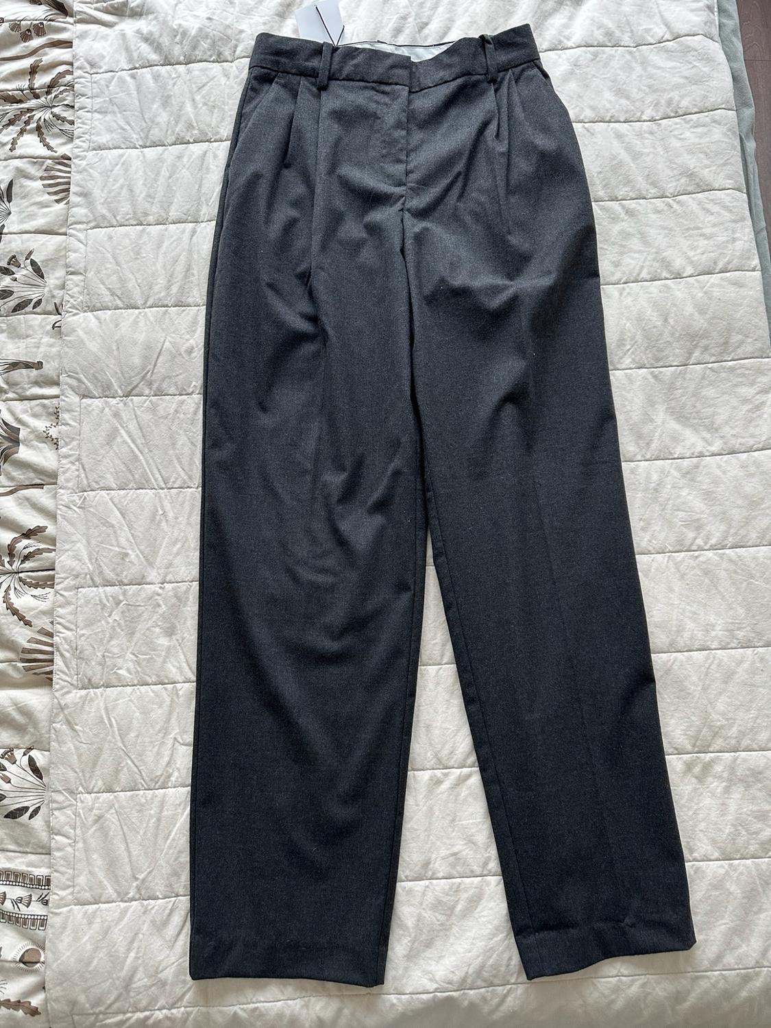 Dark grey trousers with pleats and a label, laid out on a light-coloured quilted bedspread with a pattern along the left edge.