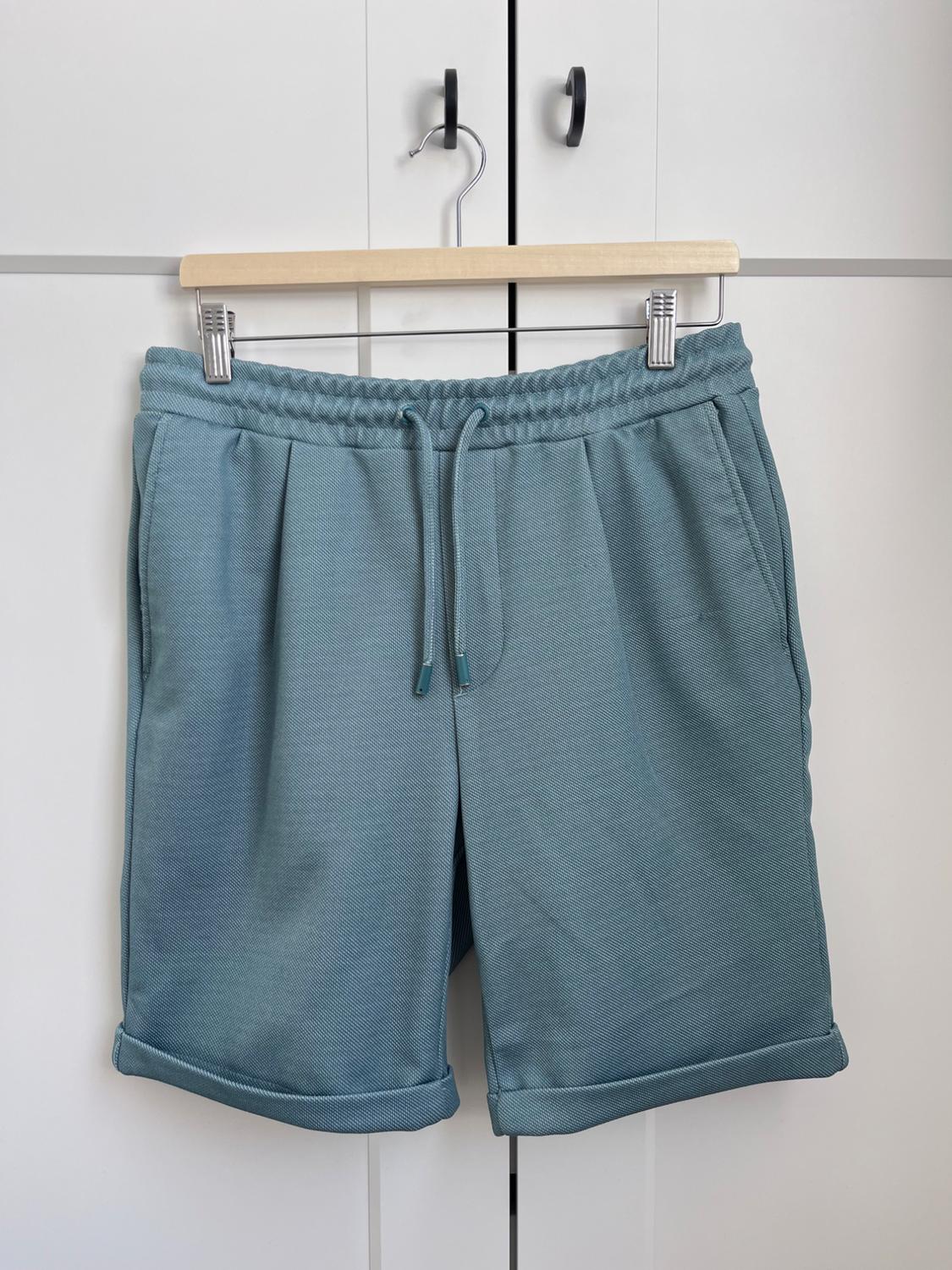 Grey-blue shorts hanging on a wooden hanger in front of a white wardrobe with black handles.